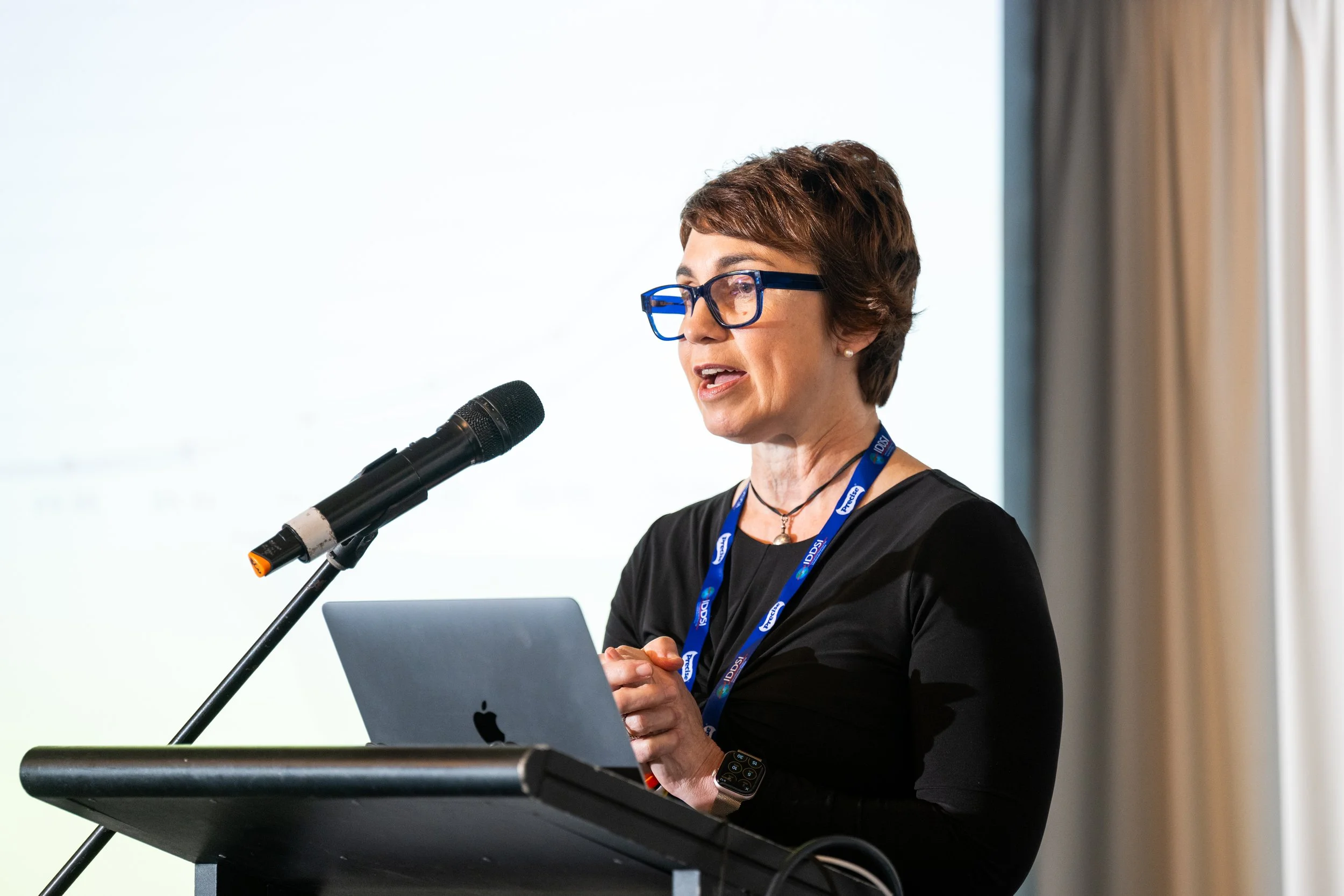 Dr Julie Cichero presenting at a conference, speaking into a microphone at a podium, with her laptop open in front of her.