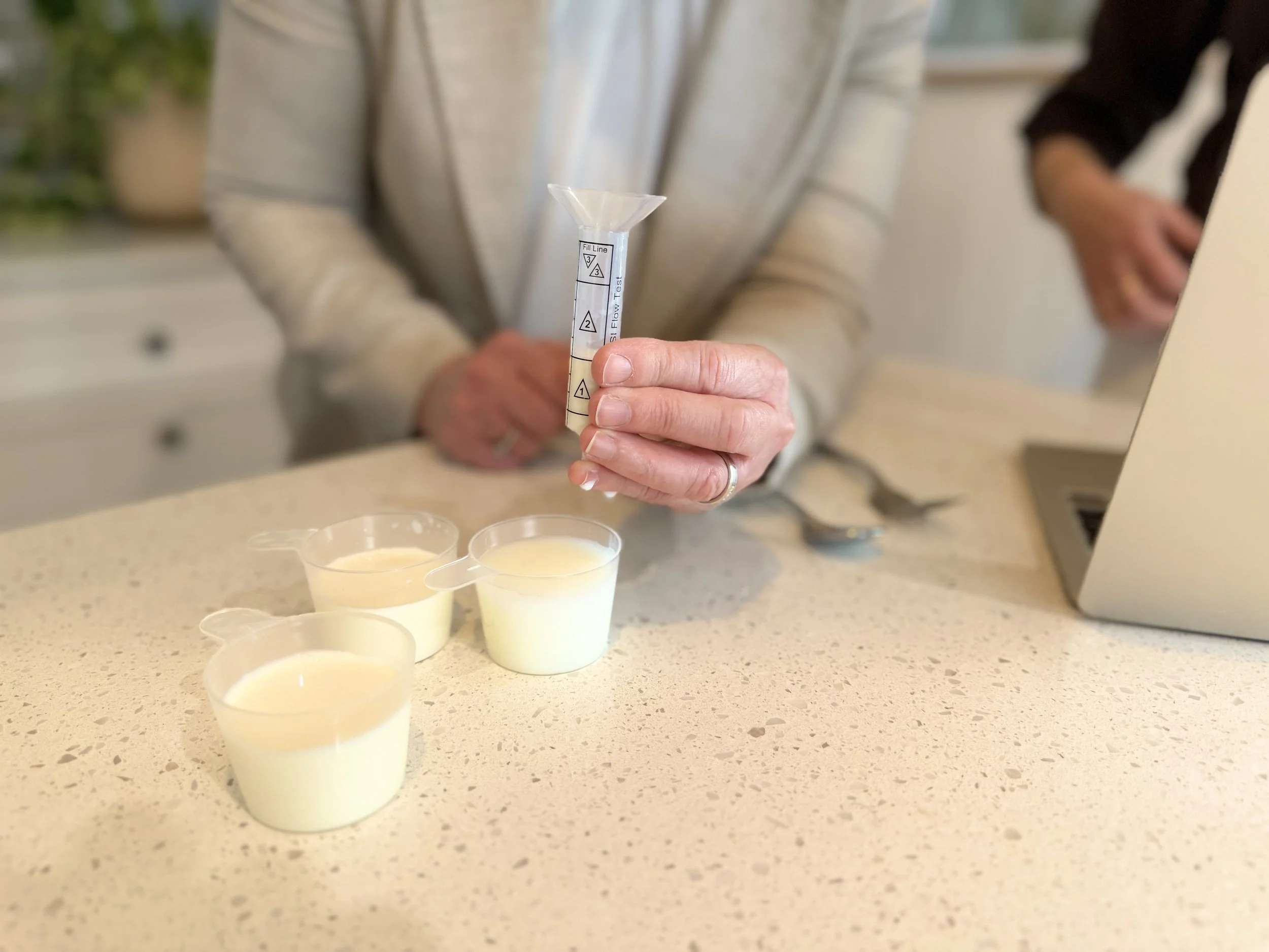 The image shows a person holding a small measuring device with a funnel on top, with several small cups of a light-colored beverage or liquid on a beige countertop in front of them. The person's face is not visible.