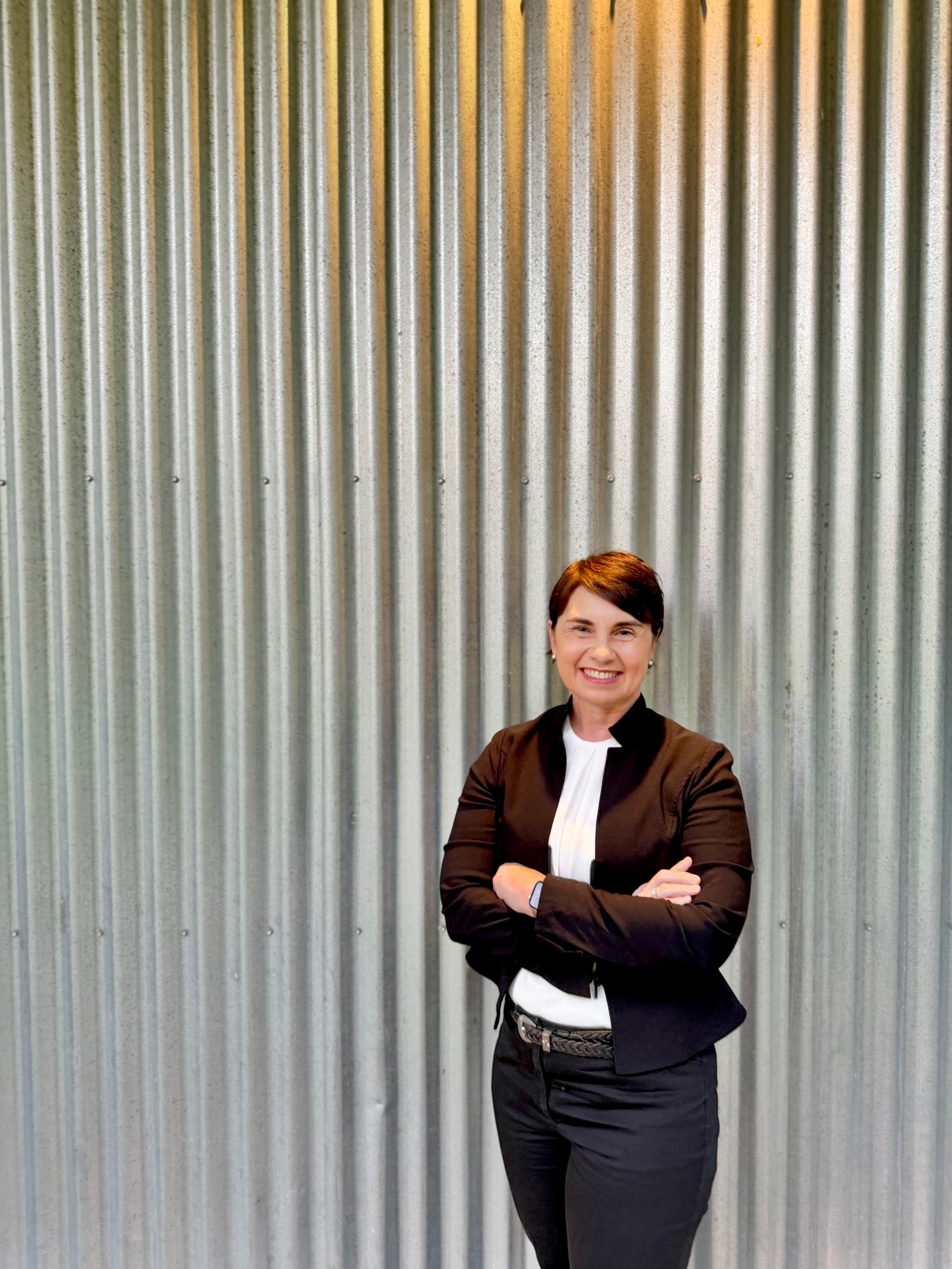 Dr Julie Cichero, co-founder of the IDDSI framework, standing with crossed arms in front of a corrugated metal wall, smiling at the camera.