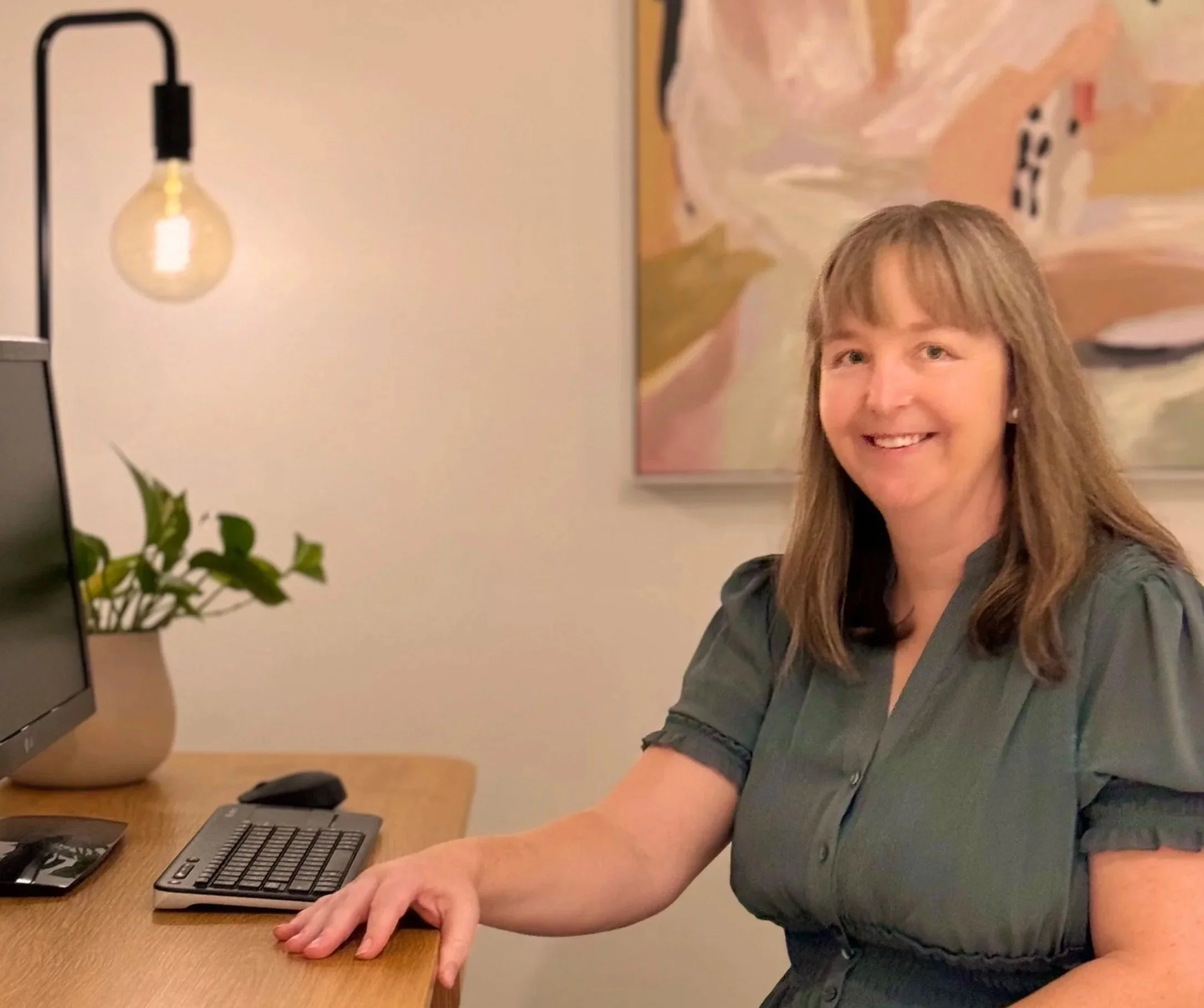 A woman with light brown hair, wearing a gray shirt, is sitting at a wooden desk with a computer keyboard and mouse. She is smiling and looking at the camera. There is a small potted plant and a framed picture on the wall behind her. A black lamp with an exposed light bulb is on the left side of the image.