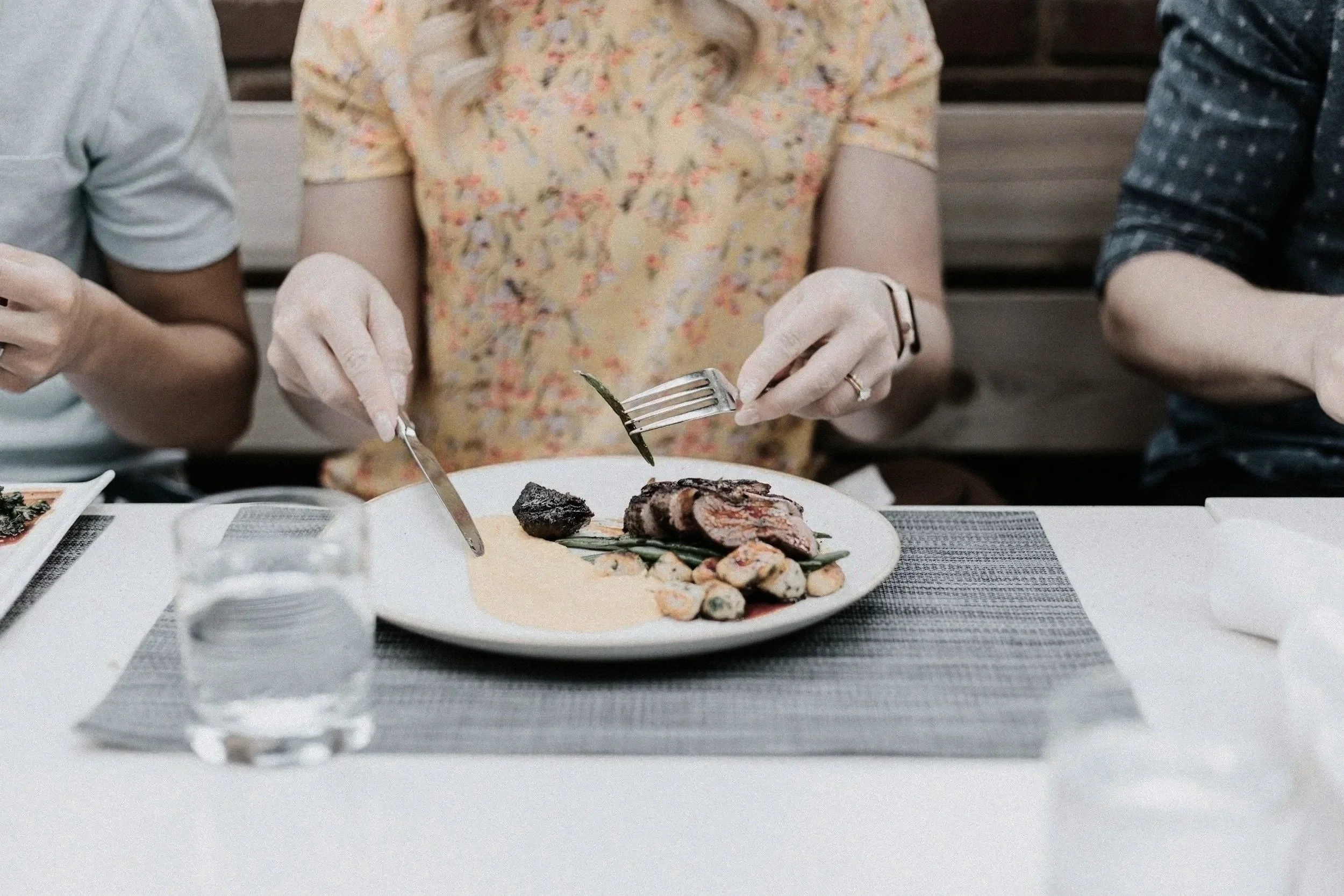 People enjoying a meal together at a table.  The woman is eating a plate of food including steak and vegetables. She has a glass of water.