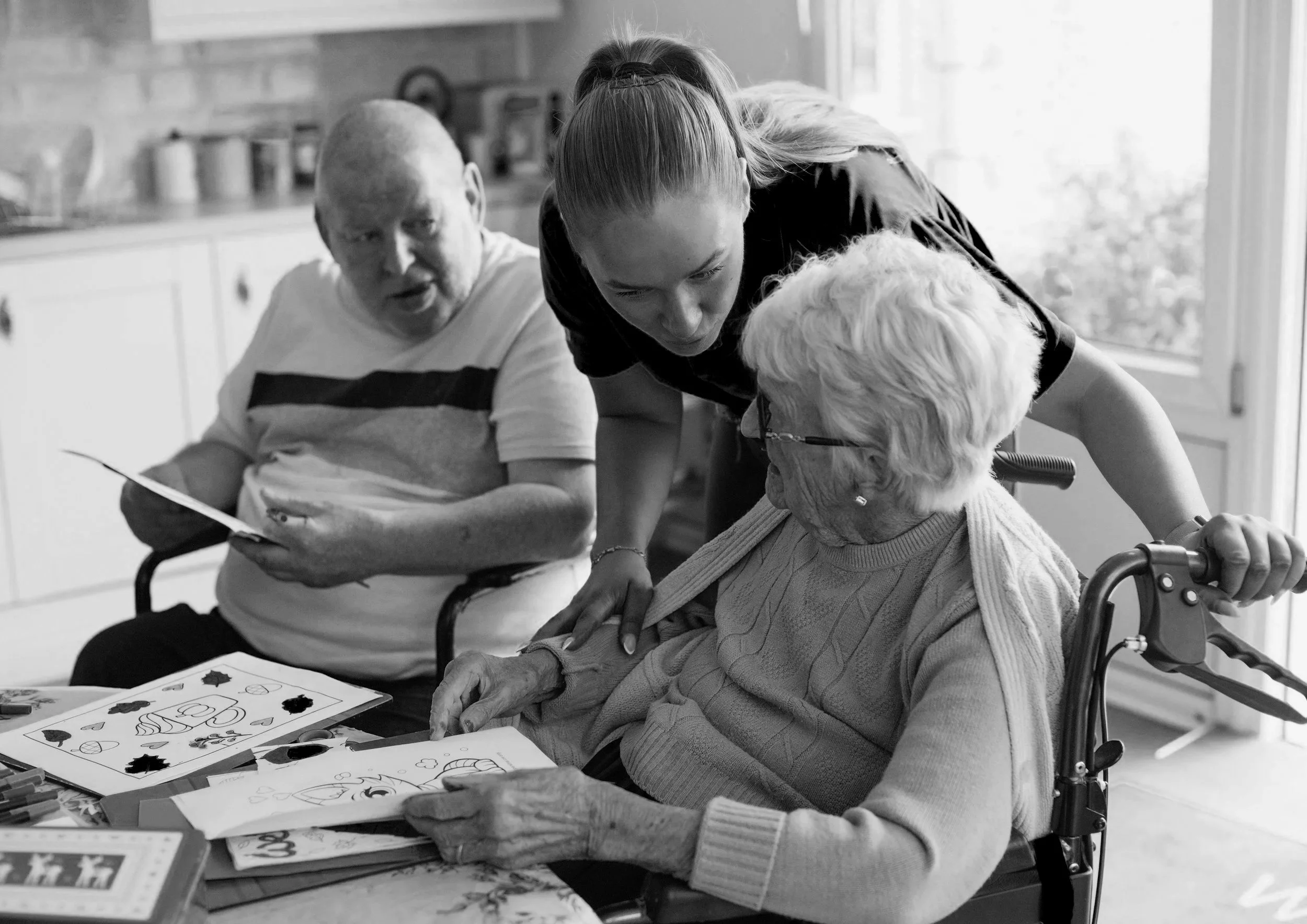 An elderly couple in wheelchairs in an aged care facility being cared for by a nurse.