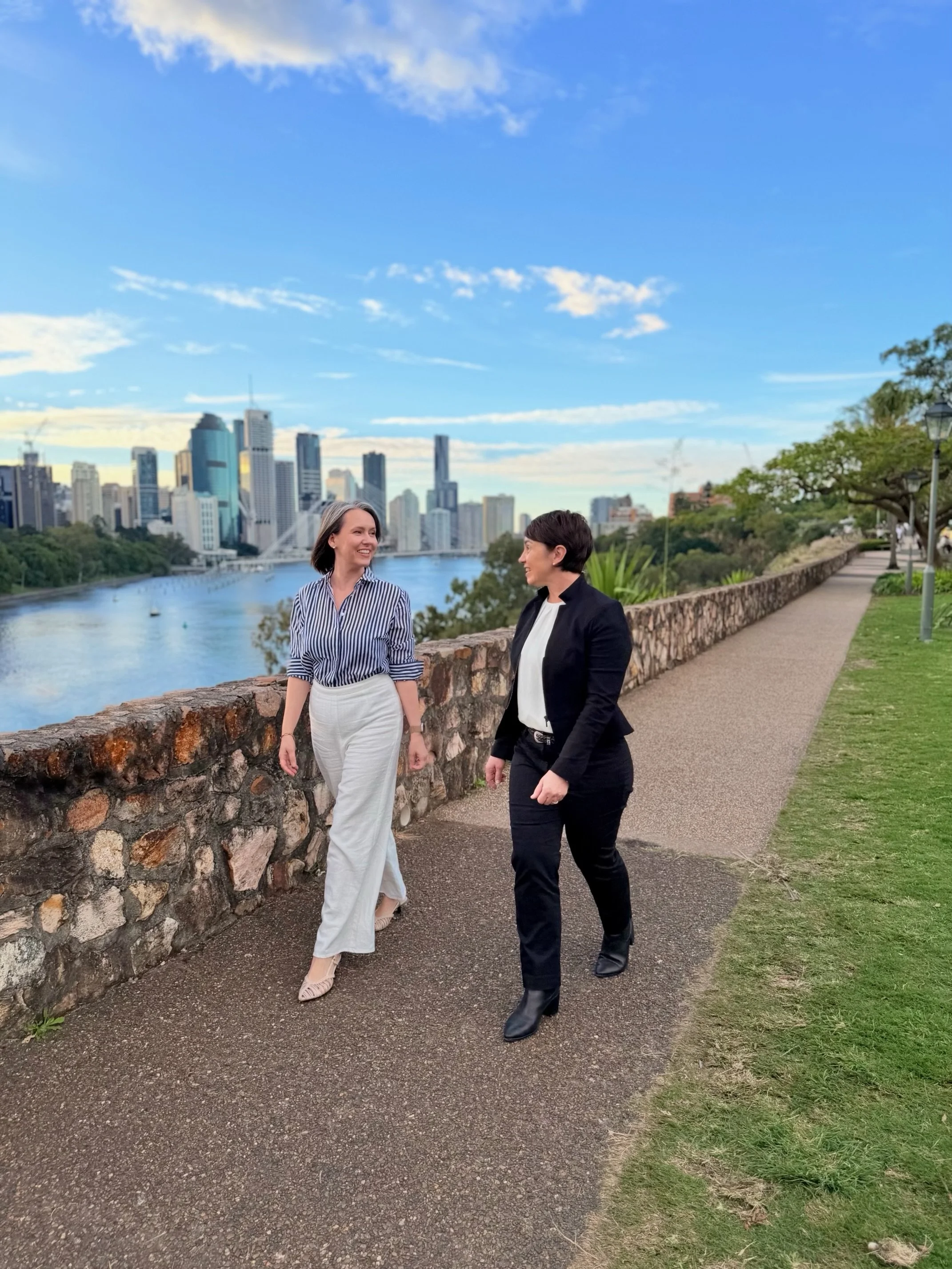 Karrie Jardine and Dr Julie Cichero walking and talking along a riverside path with the Brisbane city skyline in the background on a clear day.