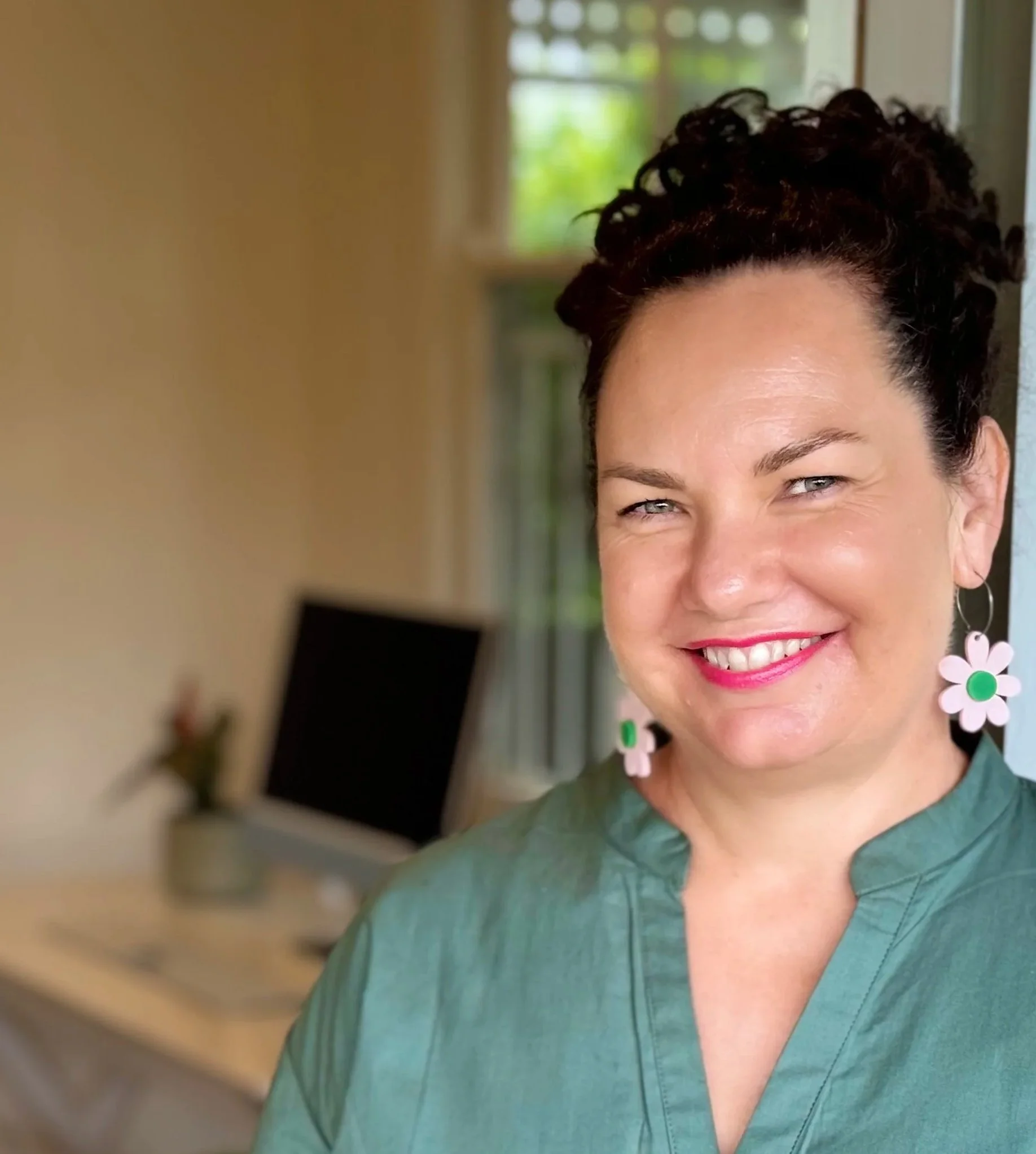 A woman with earrings smiling, wearing a teal top, and standing indoors near a window with green trees visible outside.