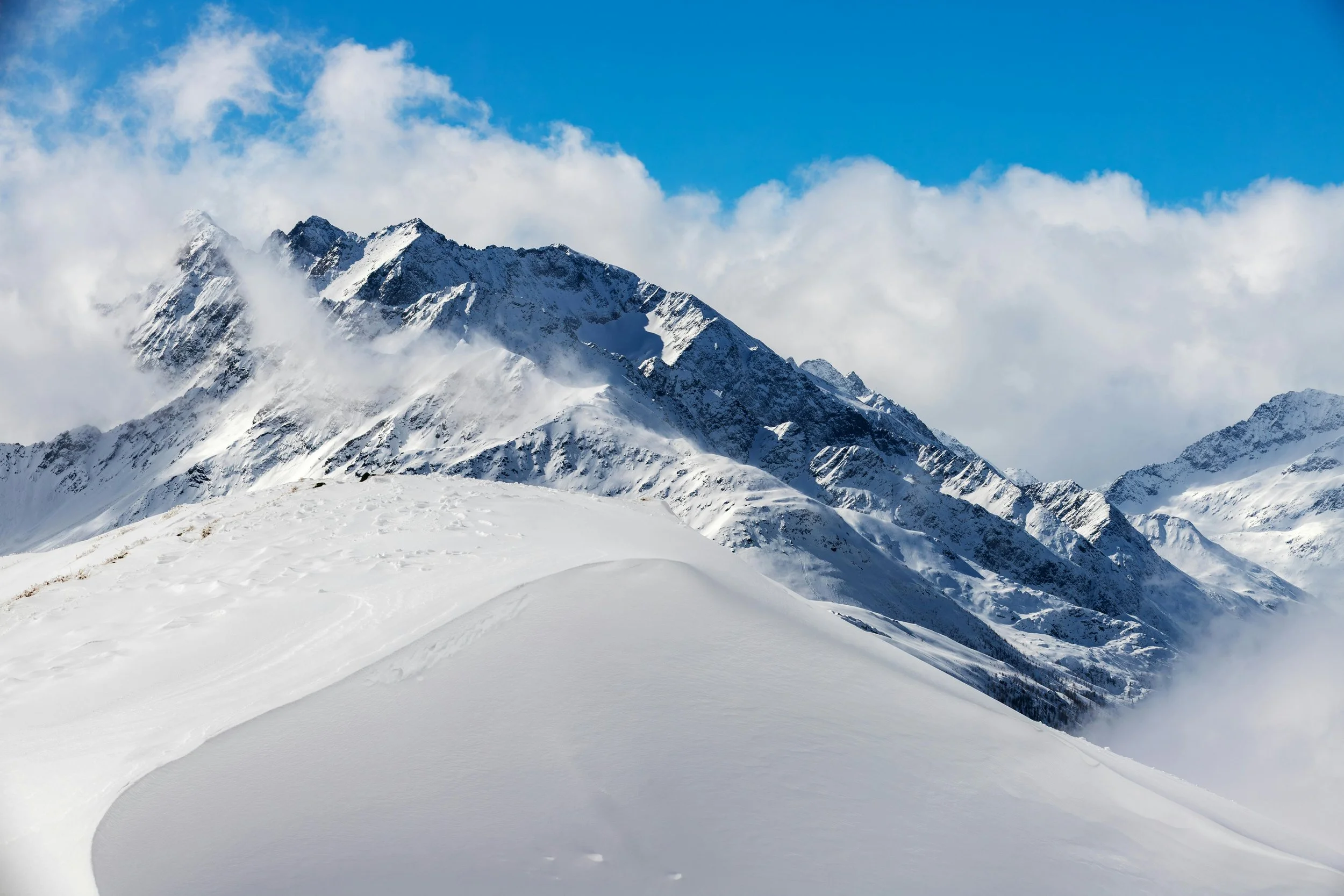 Snow-covered mountain landscape with rugged peaks, cloudy sky, and a smooth snowfield in the foreground.