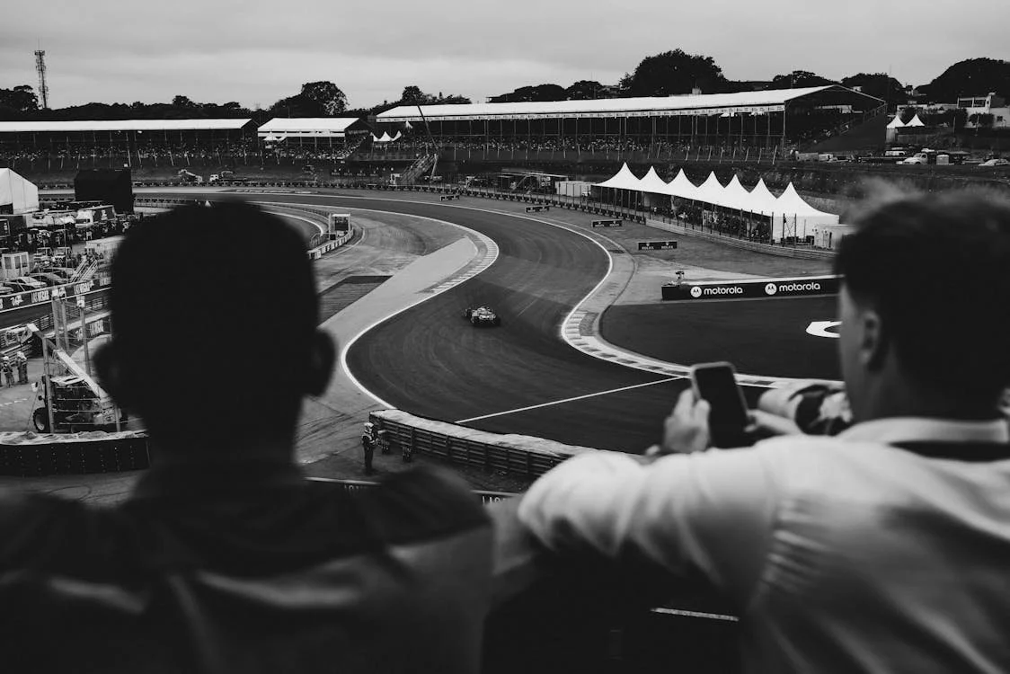 Two people watching a race track, with one person taking a photo on a smartphone, in a large outdoor motorsport event setting.