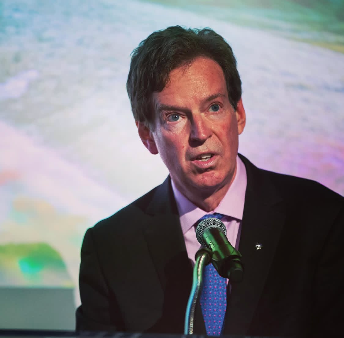A man speaking into a microphone at a conference or event, with a colorful blurred background.