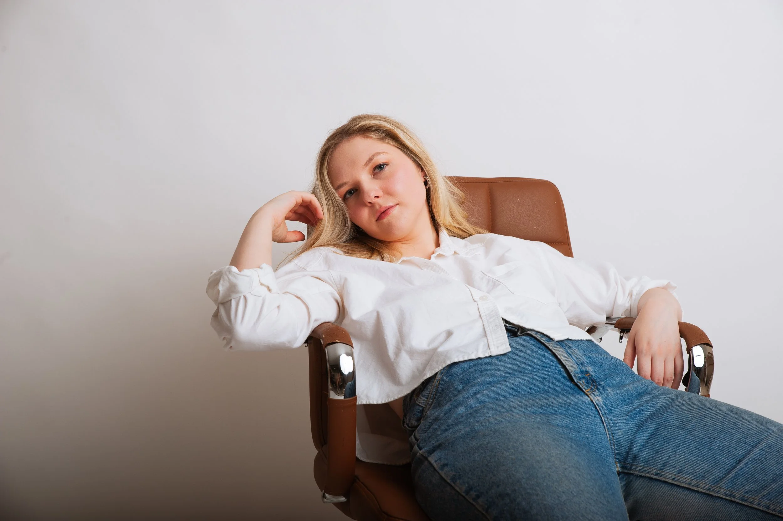 A young woman with blonde hair and a white shirt reclining in a brown chair against a plain white wall.