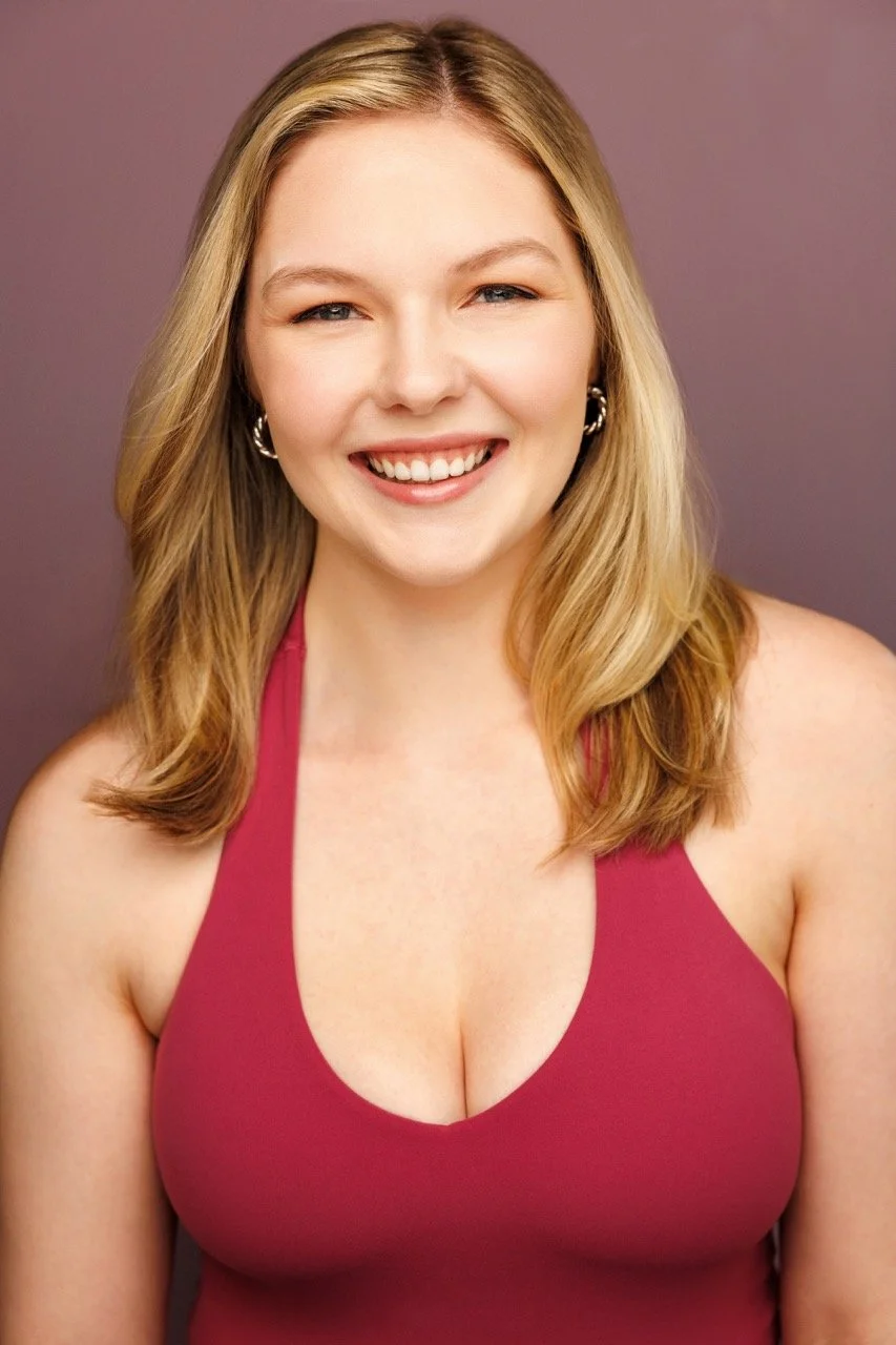 A smiling woman with blonde hair, wearing a pink sleeveless top, standing against a purple background.