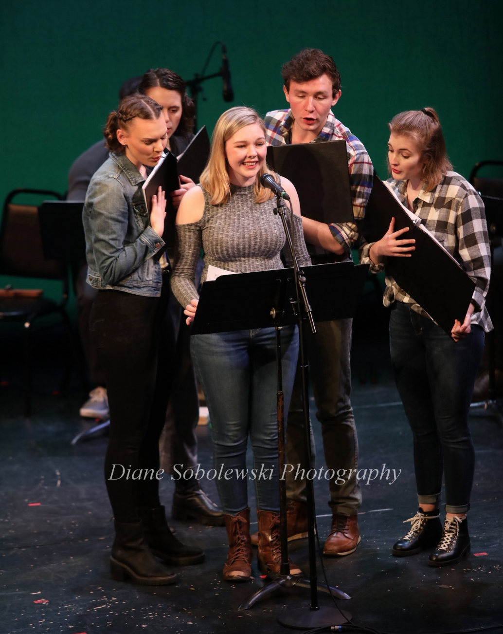A group of six young people singing together on stage, some holding music sheets, with a green backdrop and microphone stand in front.