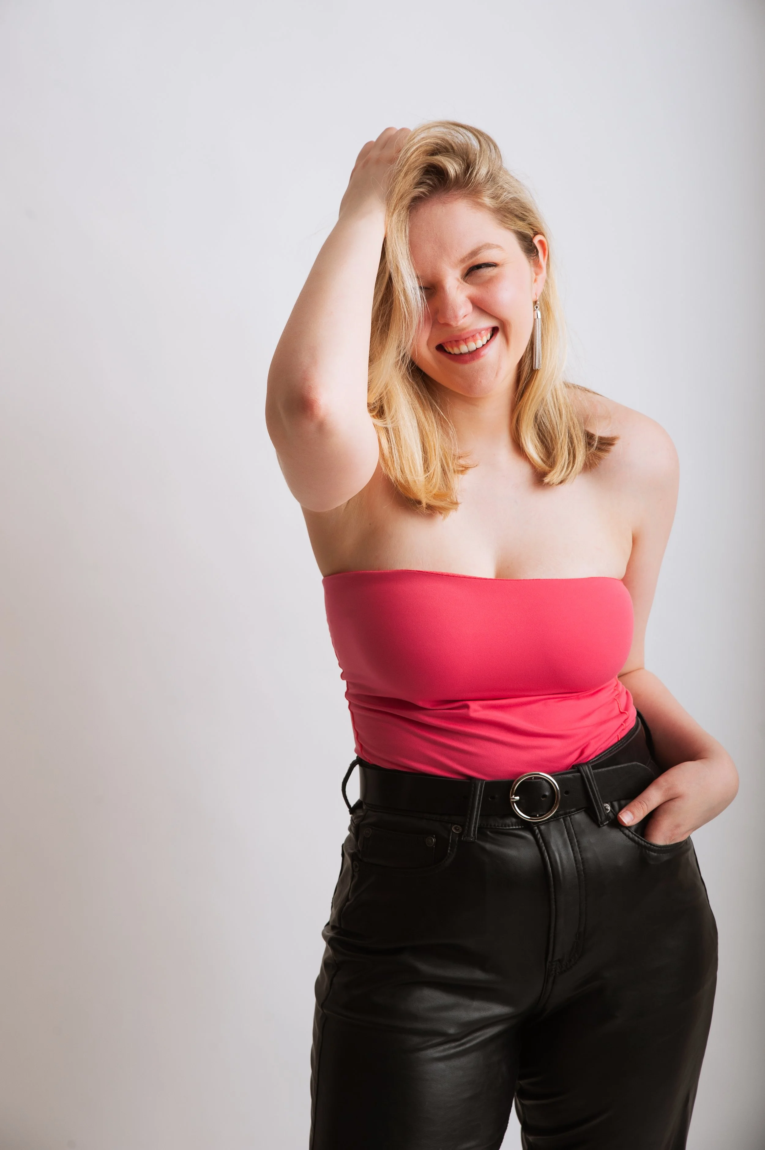A woman wearing a pink strapless top and black leather pants, smiling and posing with one hand on her head against a plain white background.