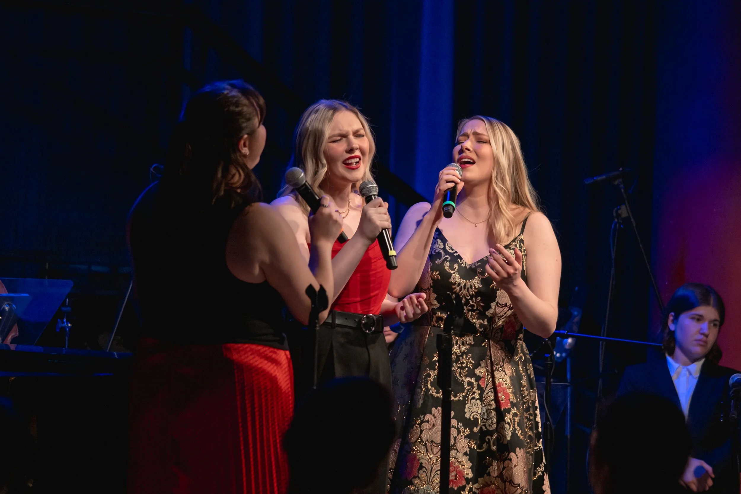 Three women singing together during a performance, holding microphones, with a blue curtain background and a set of music stands.