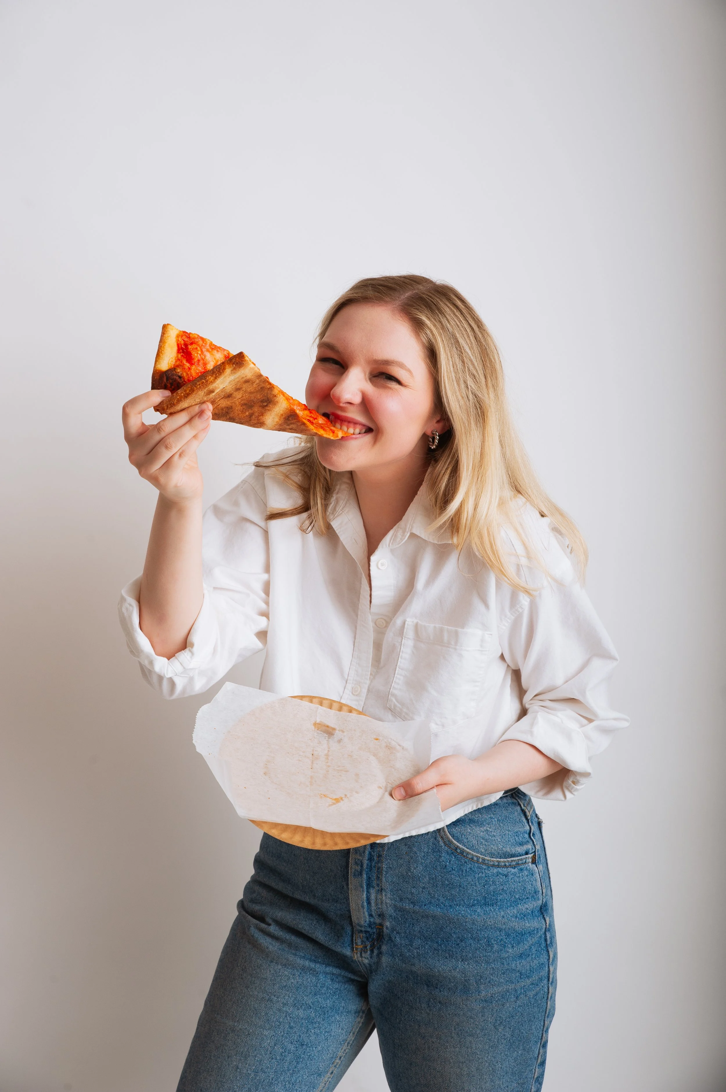 A woman with blonde hair in a white shirt and jeans enjoying a large slice of pizza, smiling with her eyes closed, against a plain light gray background.