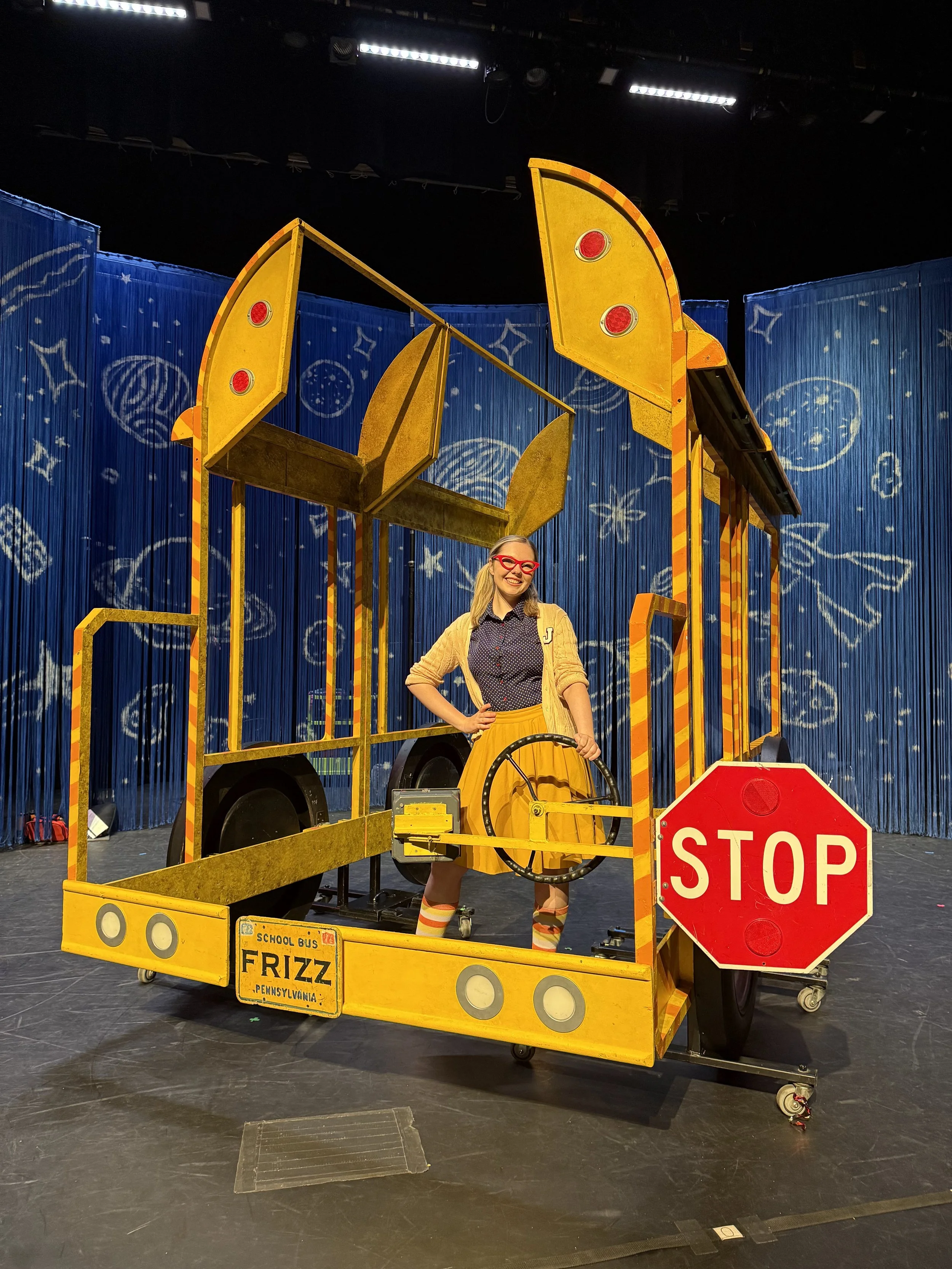 A woman dressed as a school bus driver stands on a stage behind a yellow school bus costume. The bus has a stop sign and the words "School Bus" and "Frizz" on it. The woman is smiling, wearing red glasses, a polka-dotted shirt, a yellow skirt, and a yellow cardigan. The background is a blue curtain with star and moon patterns.