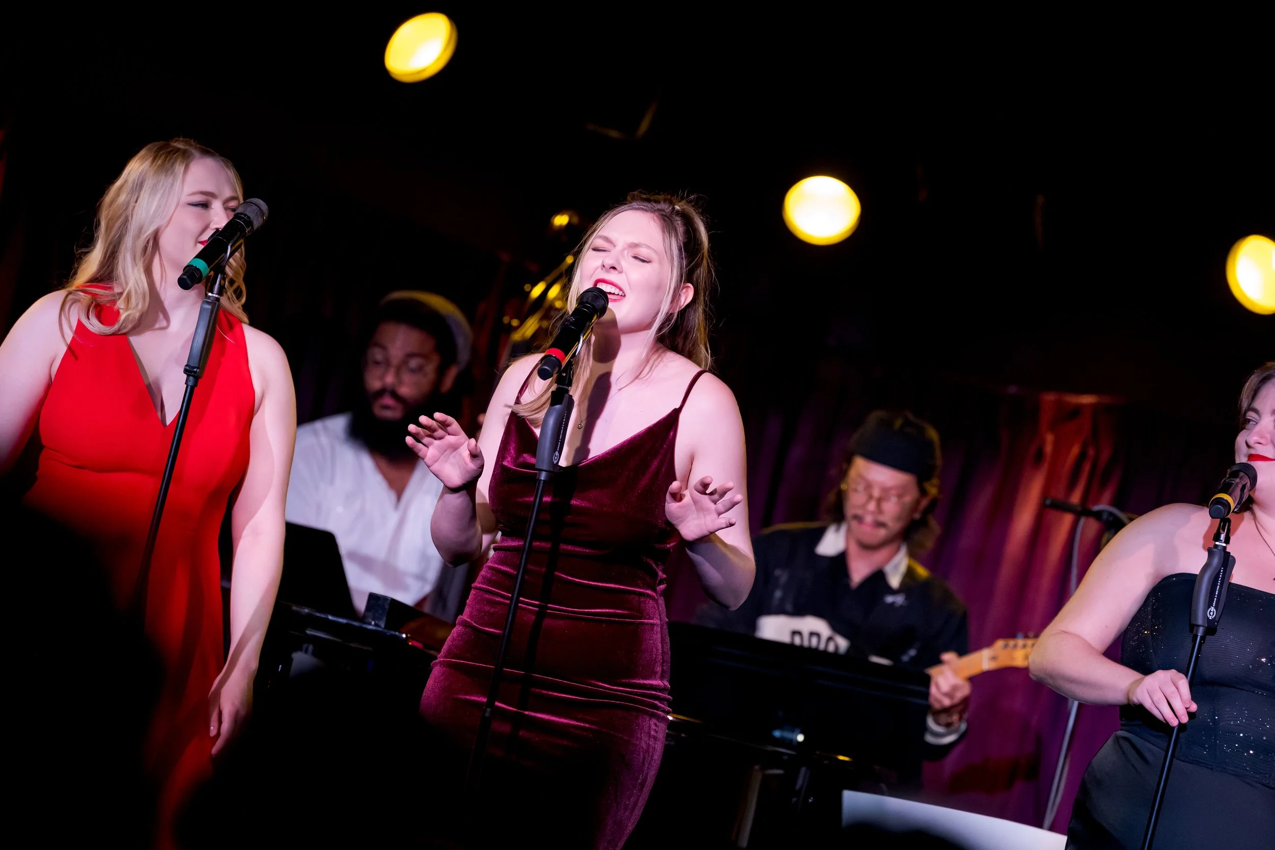A group of women singing and performing on stage, with musical instruments and background performers under warm stage lights.