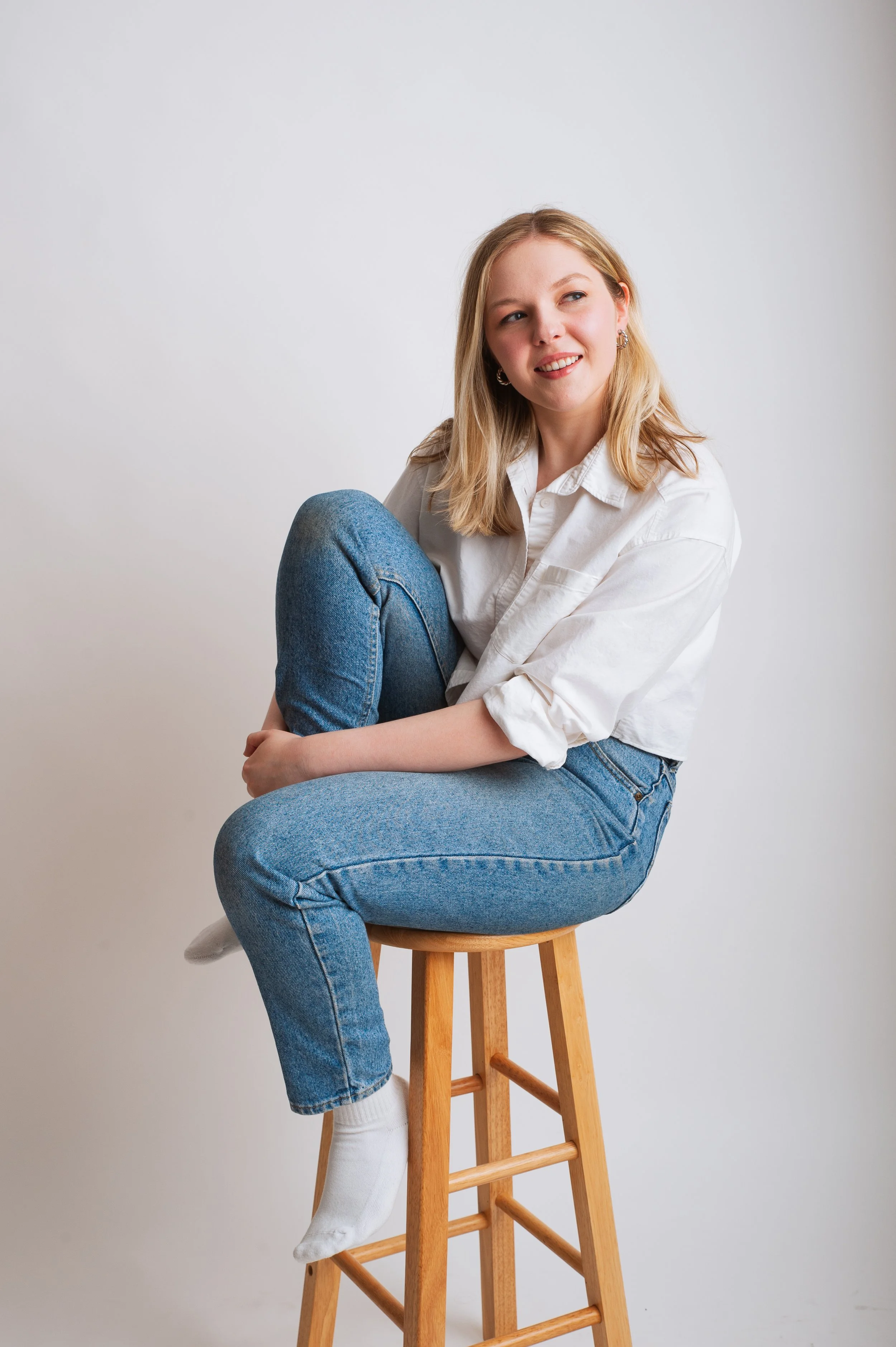 A young woman with blonde hair sitting on a wooden stool with one leg crossed over the other, wearing a white shirt, blue jeans, and white socks, against a plain white background.
