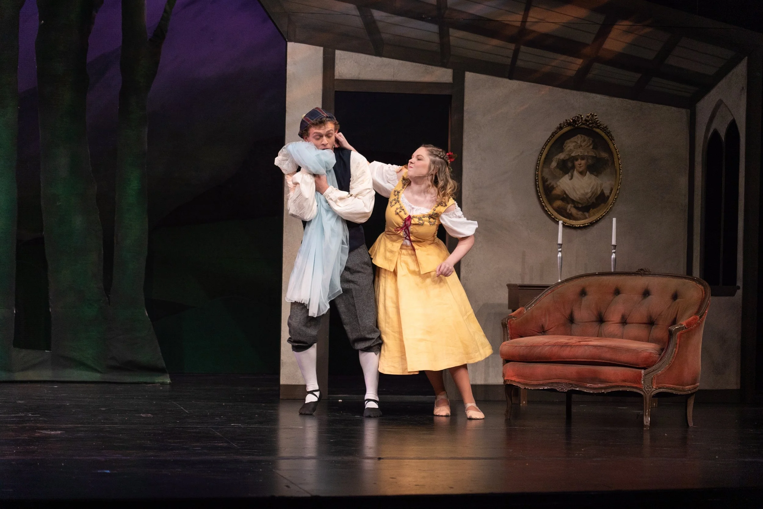 Two actors on stage performing a scene from a play. The man is dressed in historical attire and appears to be in a dramatic moment, while the woman in a yellow dress seems to be engaged with him. The background includes a portrait of a woman with a hat, candles, a sofa, and a painted set piece resembling trees.