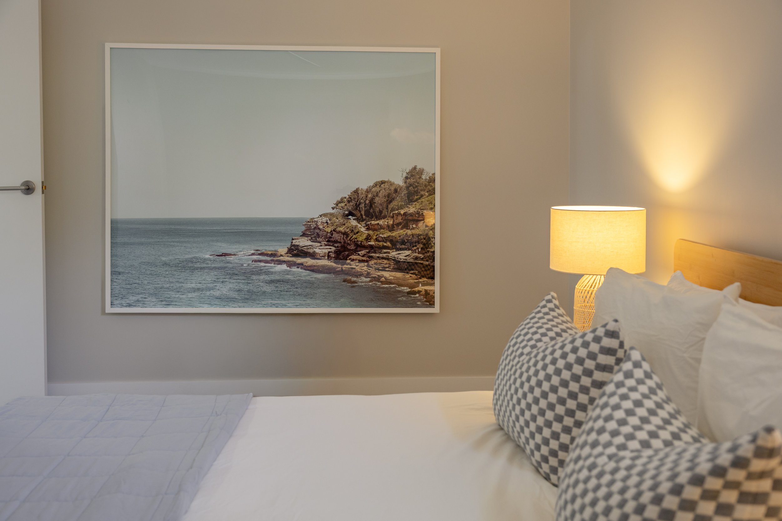A bedroom with a large window mural of a rocky coastline and ocean view, a white bed with checkered pillows, a lamp on a bedside table, and simple decor.