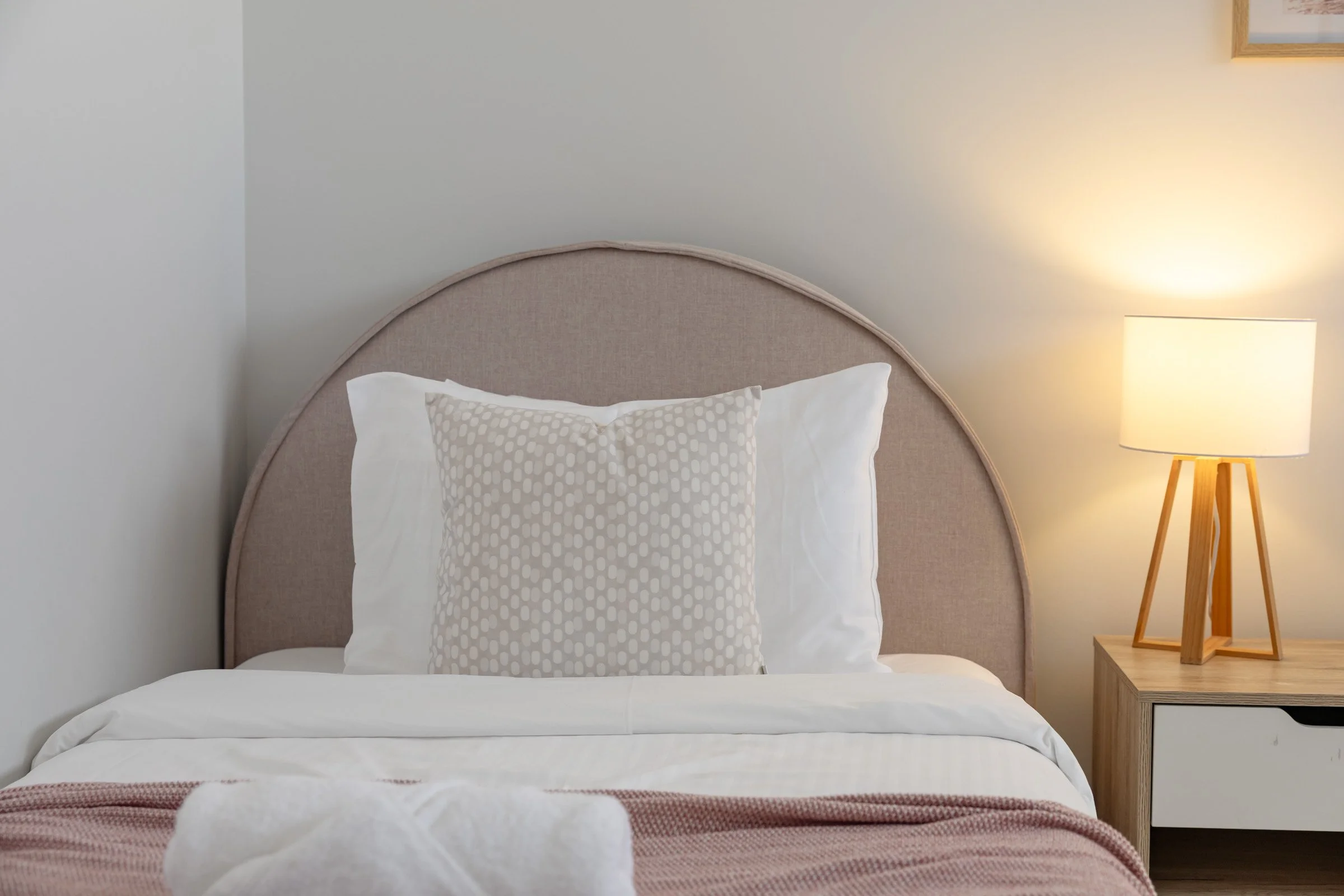 A neatly made bed with white sheets, a patterned pillow, a pink and white blanket, a beige upholstered headboard, a wooden nightstand, and a modern lamp in a bedroom with light-colored walls.