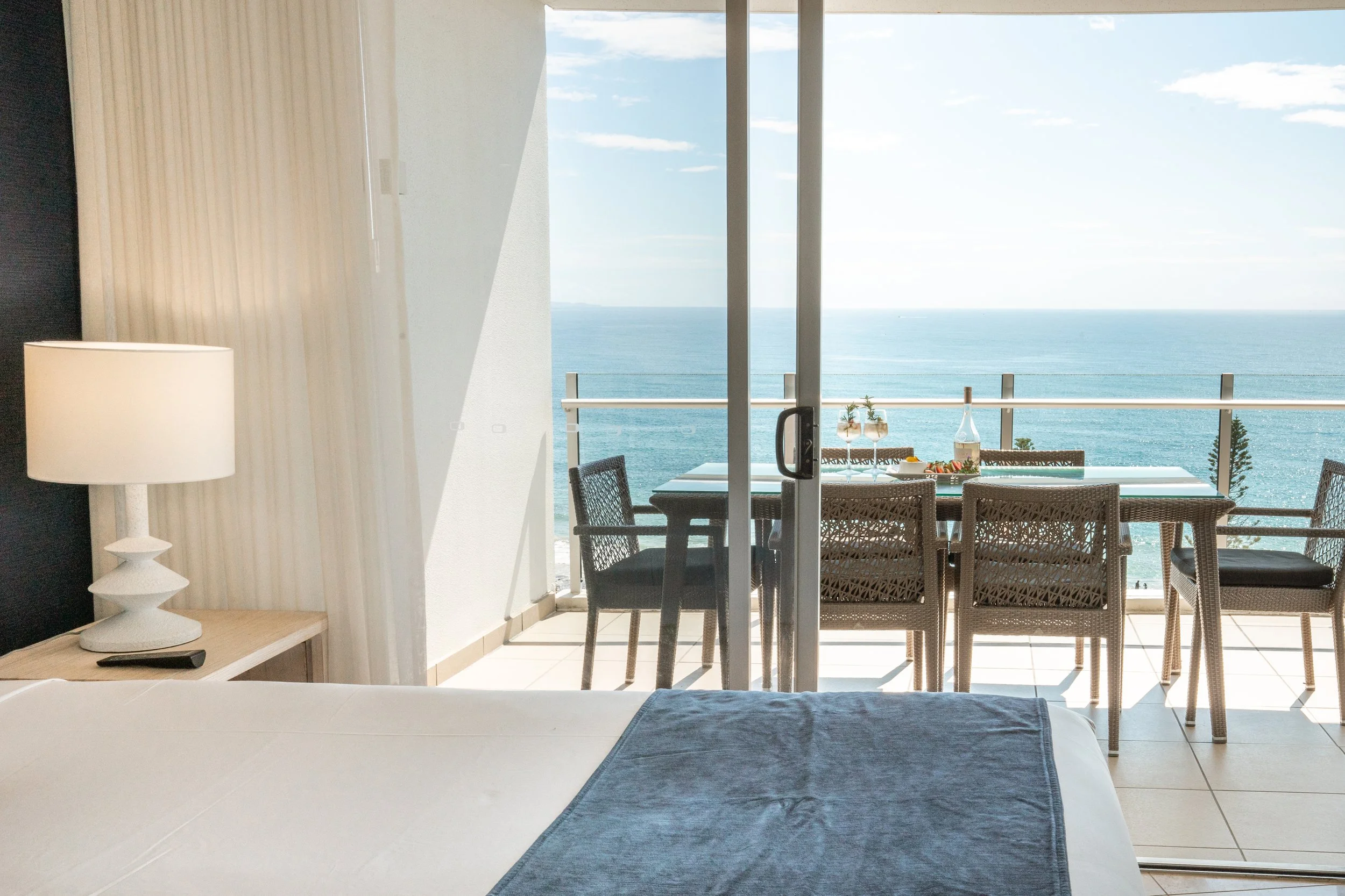 Hotel room with a balcony overlooking the ocean, with a table and four chairs, and a bottle of wine with glasses on the table.
