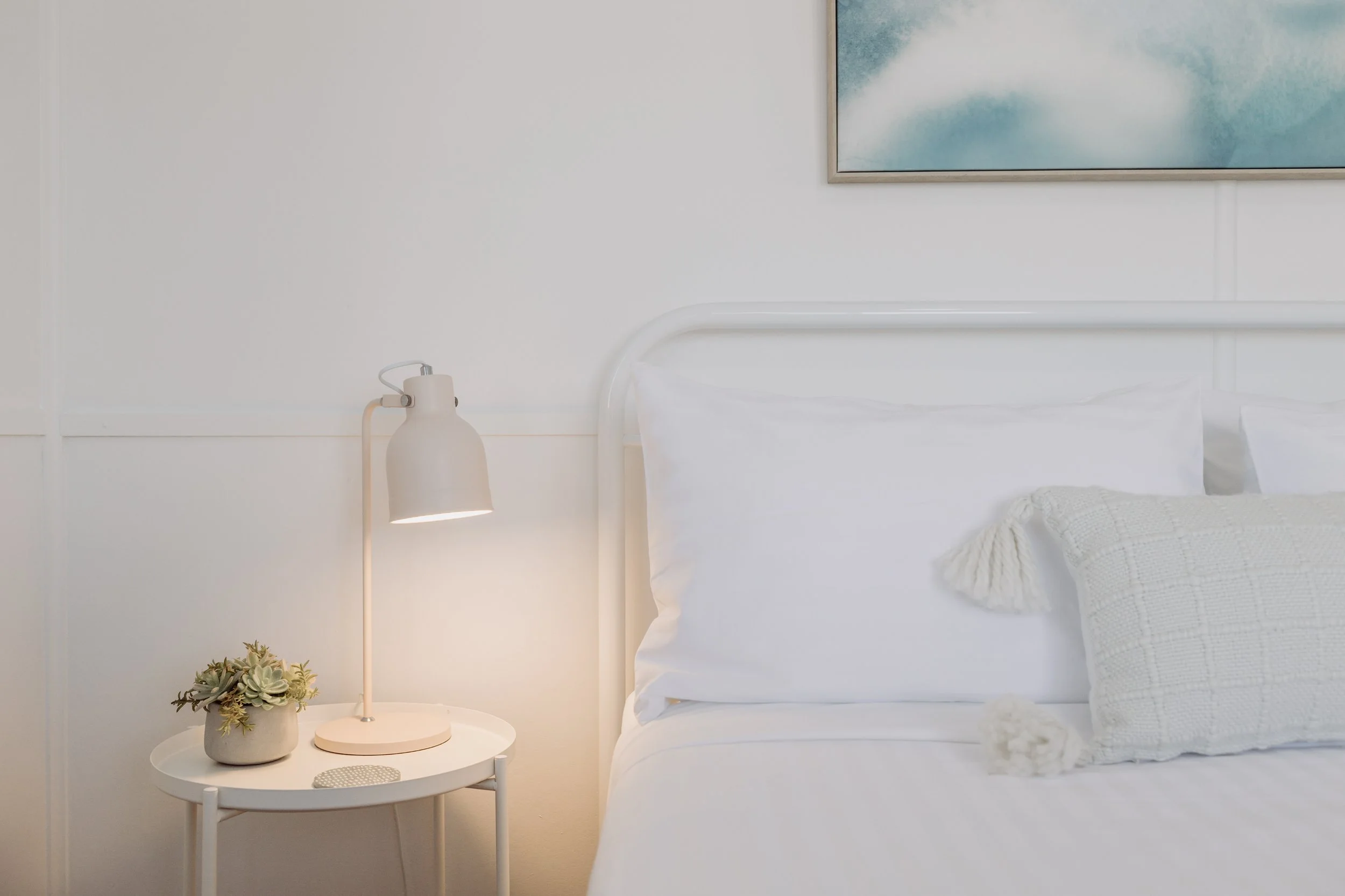 A neatly made bed with white sheets, decorative pillows, and a woven throw blanket. A straw hat rests on the bed. A wooden nightstand with a lamp and a large beach scene painting hang on the wall. The room has sheer white curtains and sheer drapes allowing sunlight to fill the space.