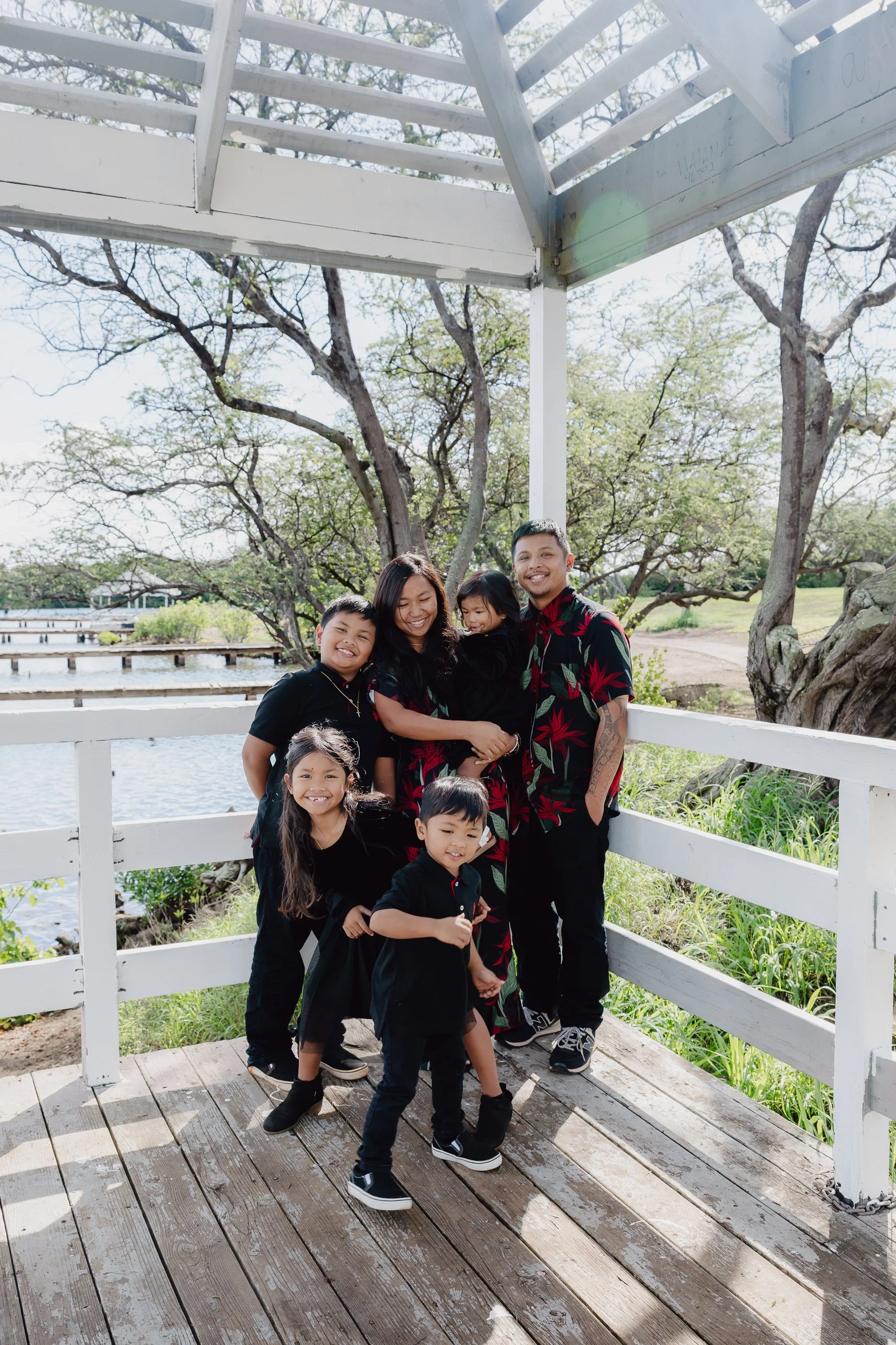 A group of seven Asian family members, including children and adults, standing on a wooden bridge outdoors near a body of water with trees in the background, smiling at the camera during daytime.