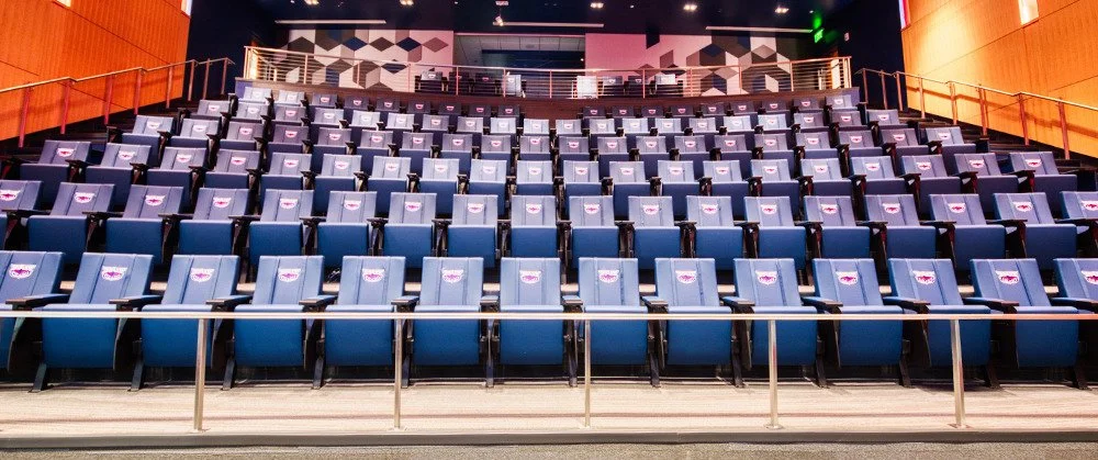 Empty blue theater seats with United Airlines logo coverings, facing a stage with dark walls and geometric patterns.