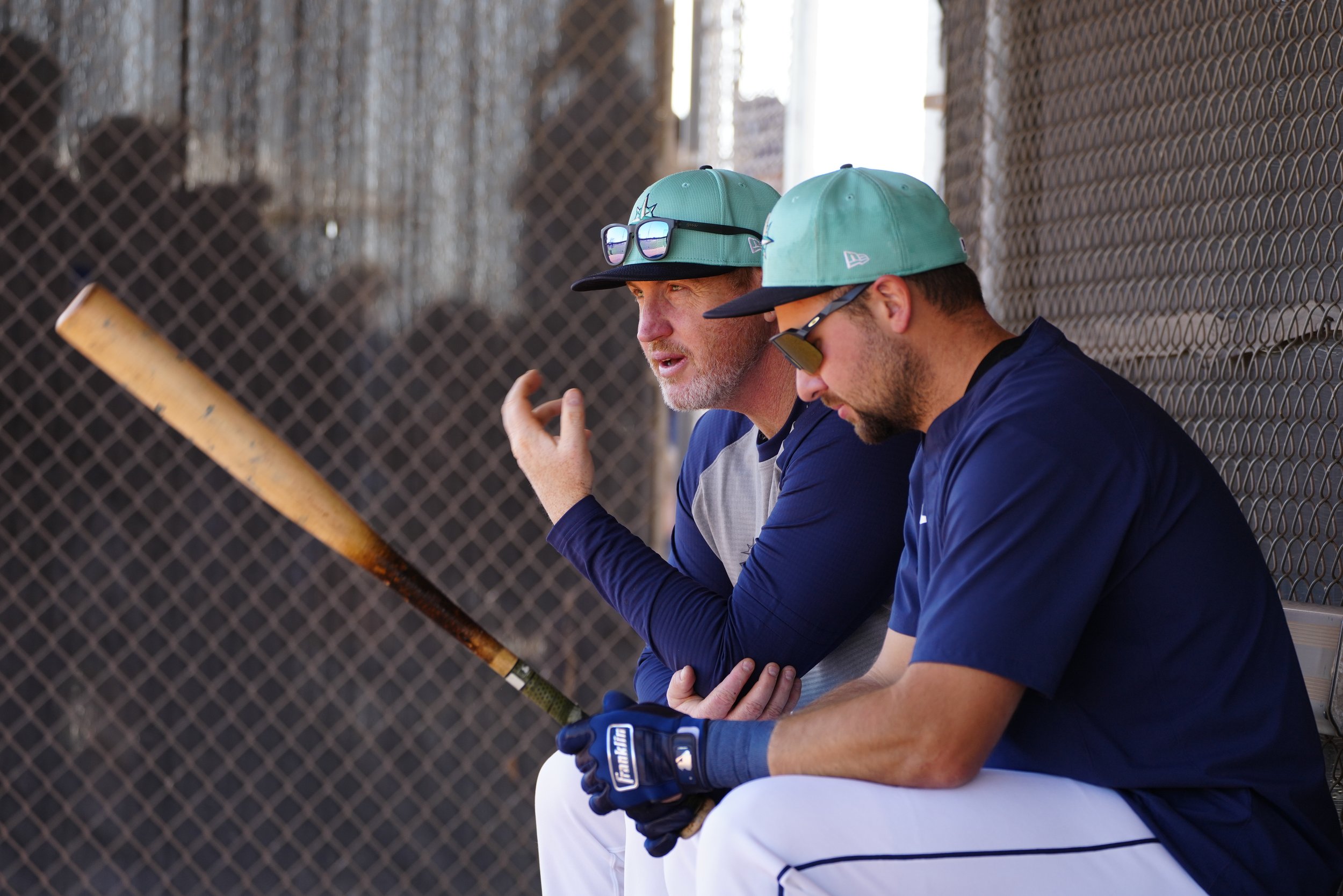 Two baseball players sitting on a bench, wearing caps and sunglasses, resting with one holding a bat and talking, against a chain-link fence background.