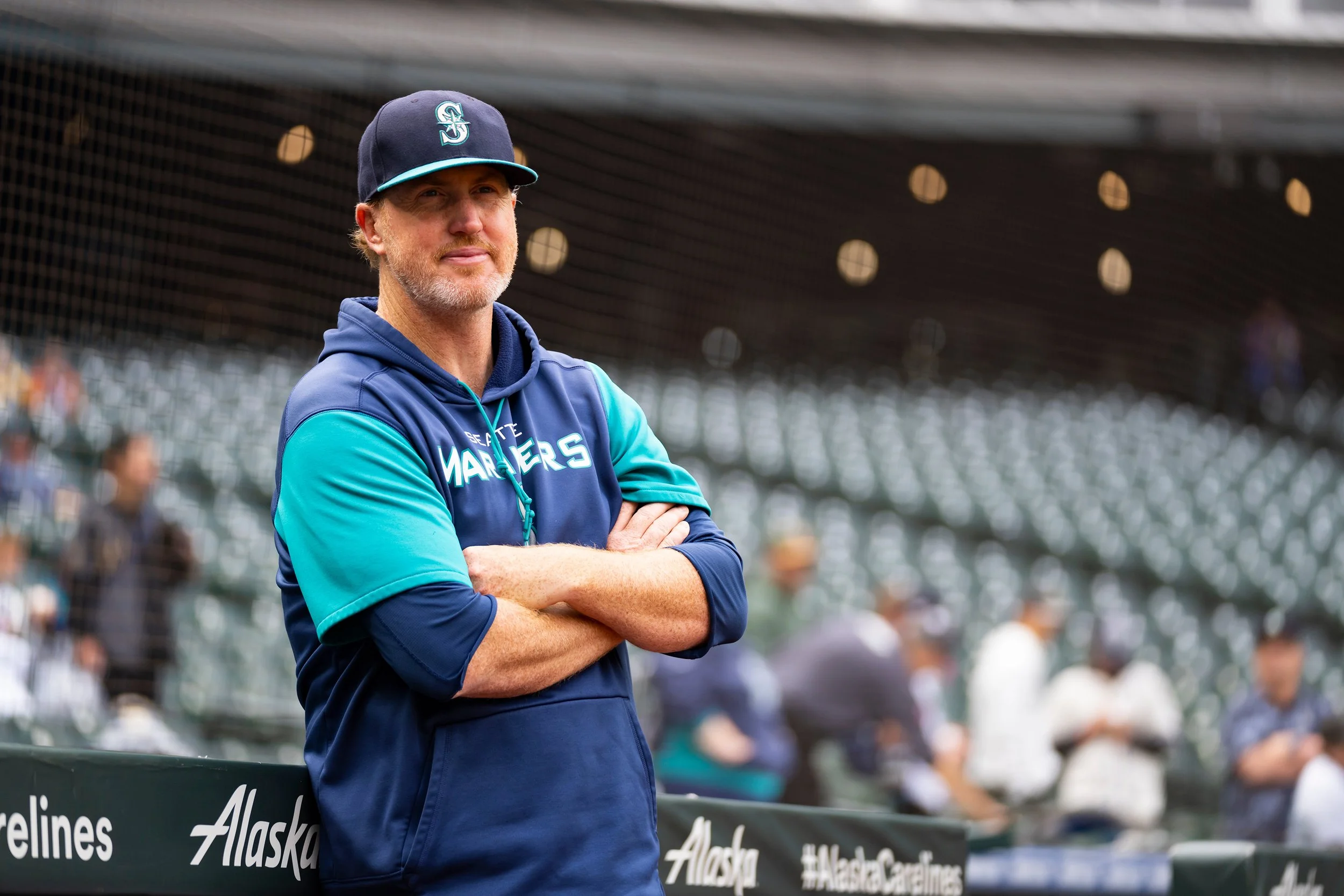 A man in a Seattle Mariners jacket and cap stands with arms crossed at a baseball game, looking attentive, with stands and spectators blurred in the background.