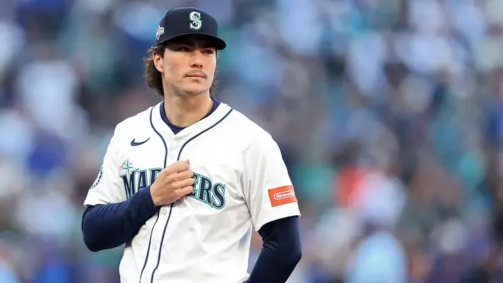 A male baseball player in a white Mariners uniform and cap standing on a baseball field.
