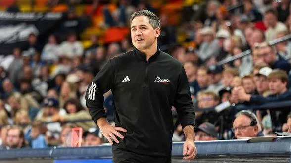 A man standing on a sports sideline with a crowd in the background, wearing a black jacket with an Adidas logo and the word 'Satellite' on it.