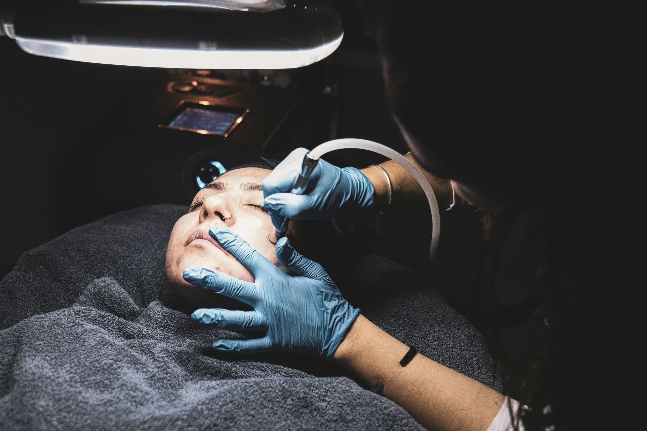 A person lying down with their eyes closed receives a facial treatment from a technician wearing blue gloves, using specialized equipment in a darkened room.