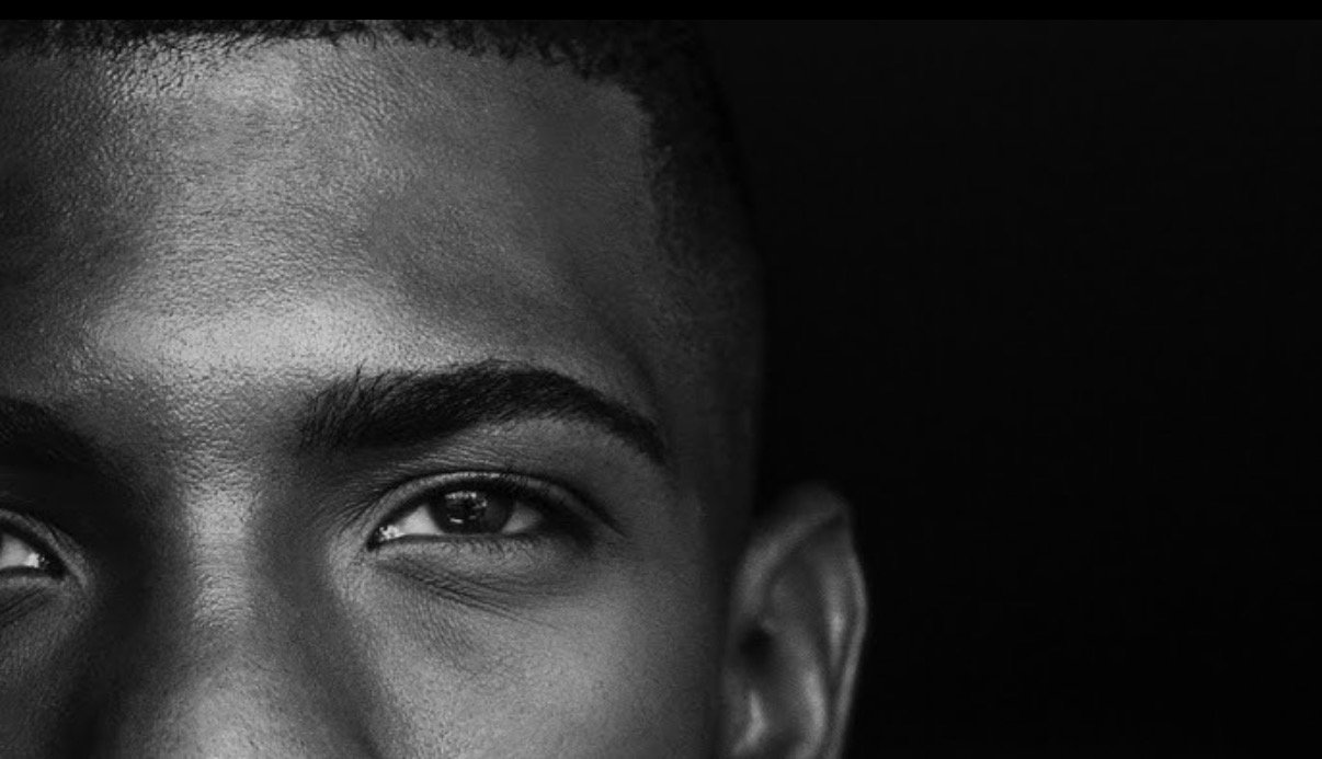 Close-up of a young man's face with a dark background, focusing on one eye, eyebrow, and part of his forehead and ear.