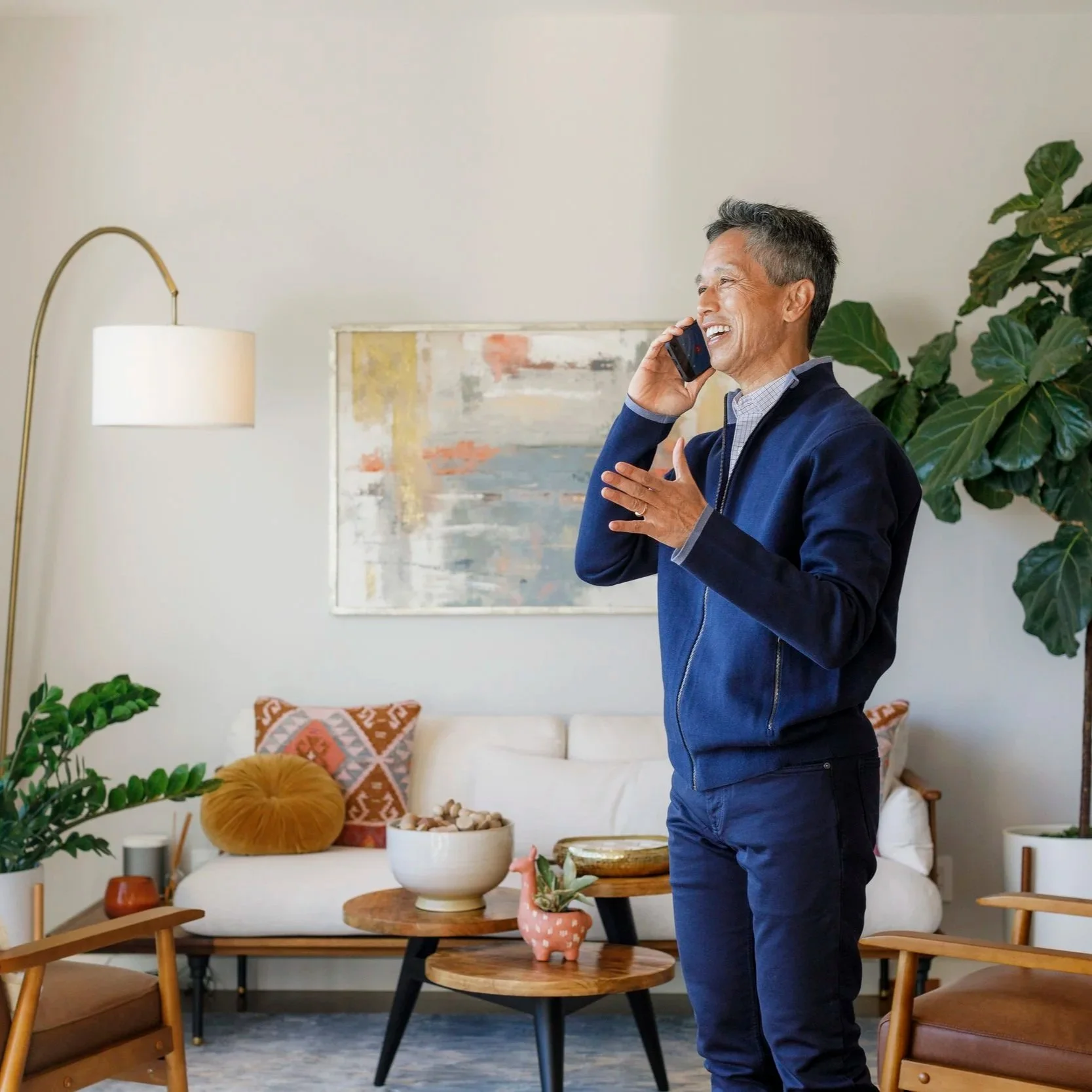 An Asian middle-aged man with gray hair smiling and talking on a mobile phone in a modern living room.