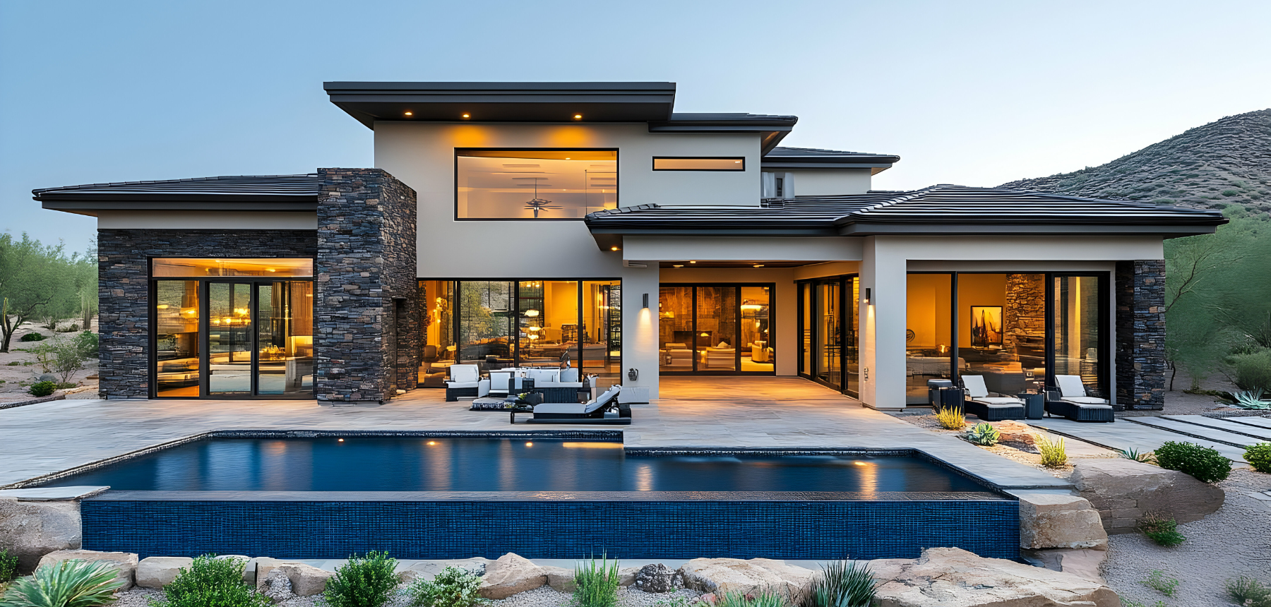Modern two-story house with stone and white exterior, large glass doors, and a swimming pool in the backyard, surrounded by desert landscape and mountains at dusk.