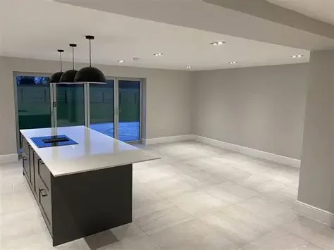 Empty modern kitchen with a white island, black pendant lights, gray walls, and large windows.