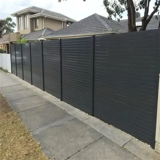 A tall black privacy fence runs alongside a sidewalk in a residential neighborhood, with houses and trees in the background.