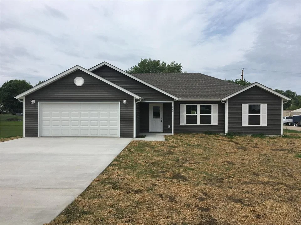 New single-story house with dark siding, white shutters, attached garage, concrete driveway, and front door, located in a suburban area.