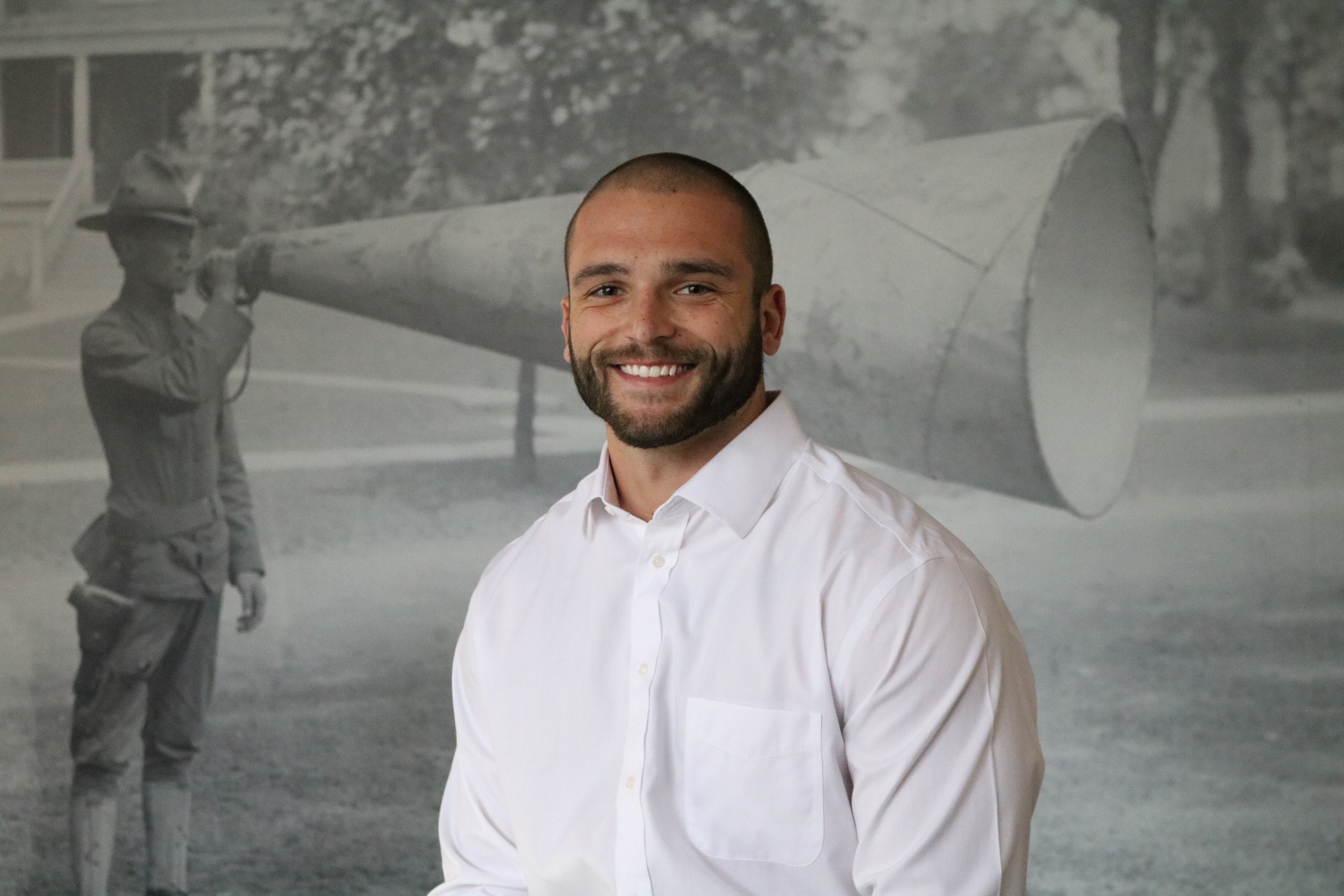 A smiling man with short hair and a beard wearing a white button-down shirt, standing in front of a historic black-and-white photograph of a person in a military uniform holding a large object, in an indoor setting.