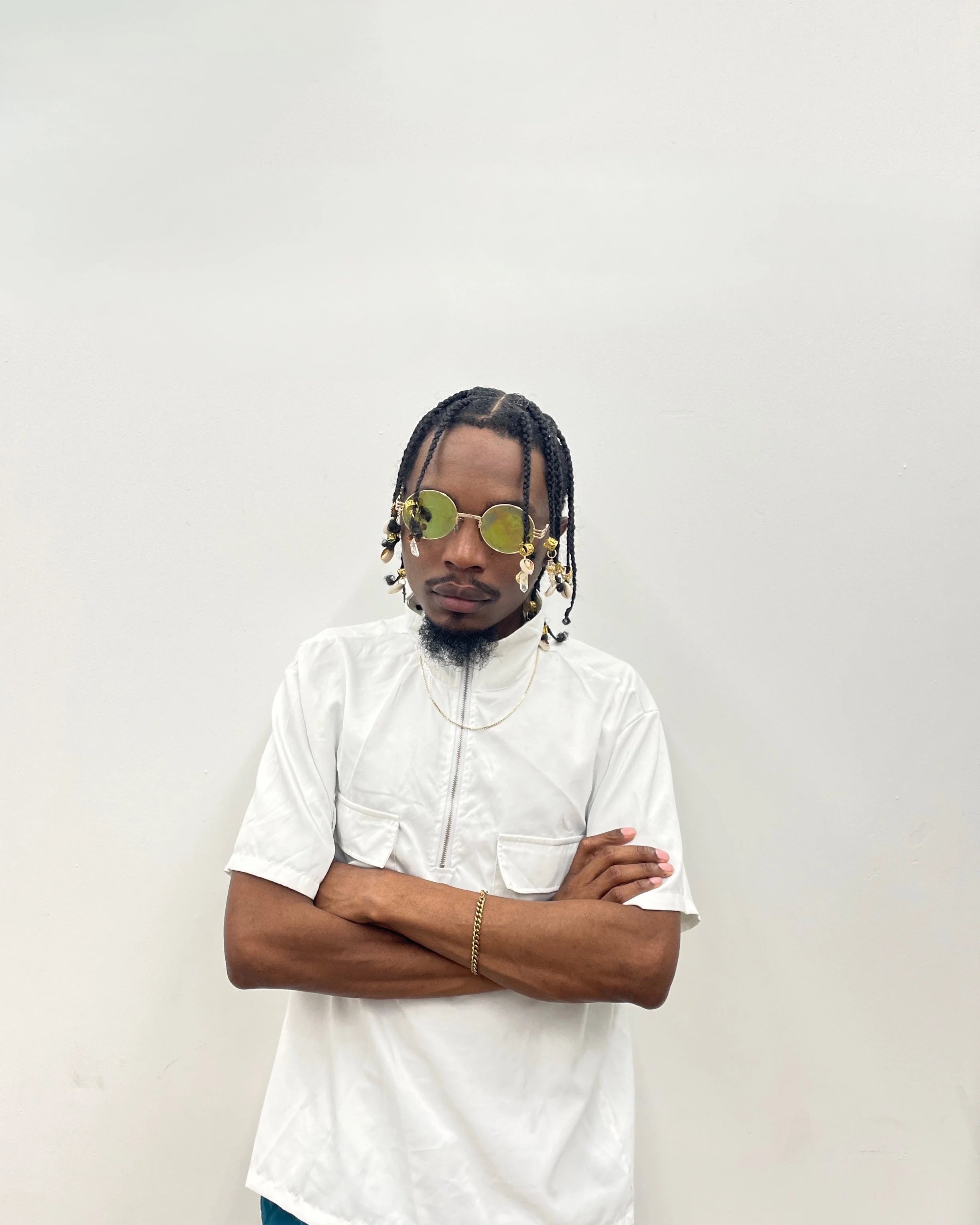 A man with braided hair wearing sunglasses, a white zip-up shirt, and gold jewelry stands against a plain white wall with his arms crossed.