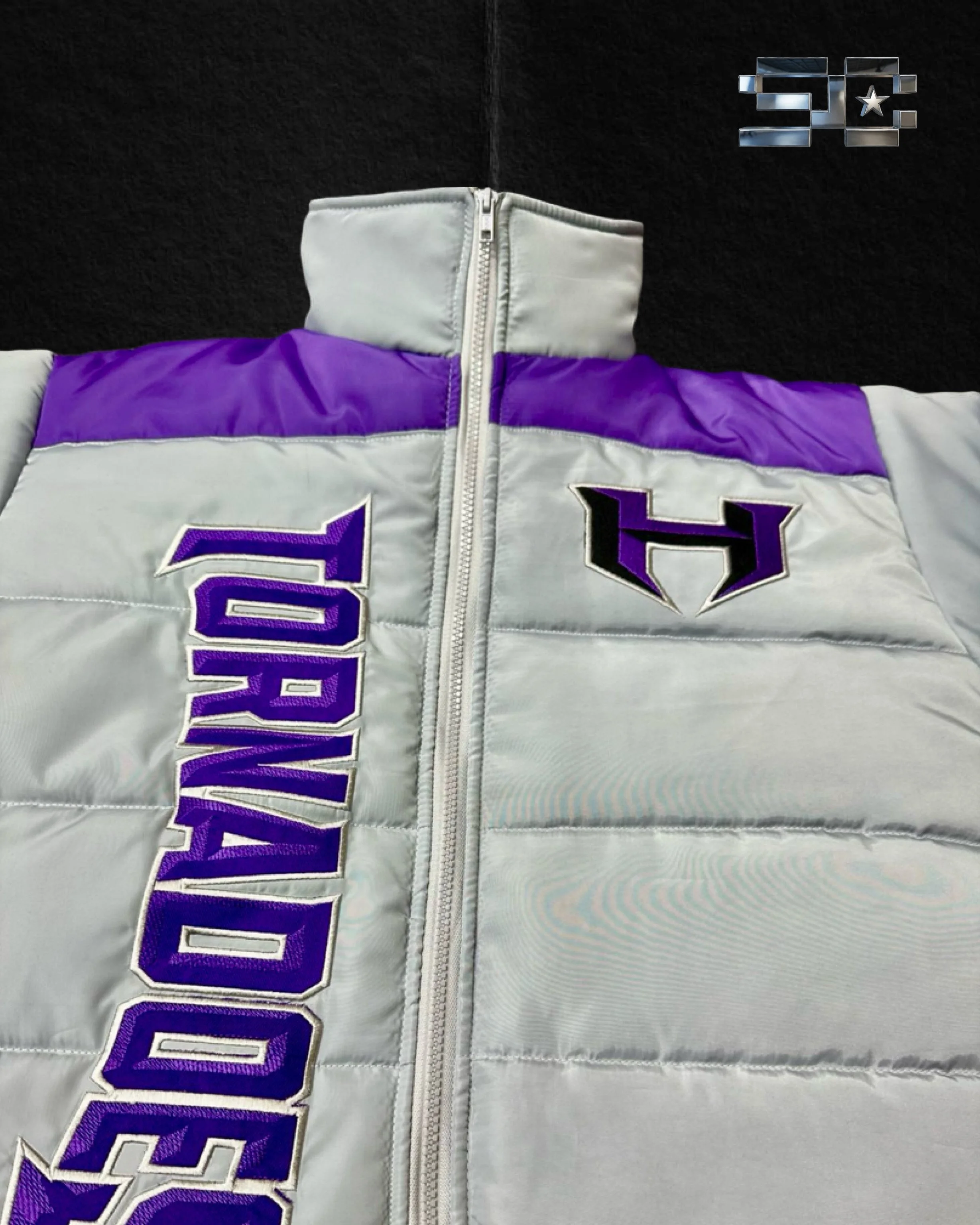 A racing jacket with a white main color, purple and black accents, and large embroidered letters spelling 'COWBOY' down the front. The jacket has a high collar, a zipper in the middle, and a black background with a star and metallic logo in the top right corner.