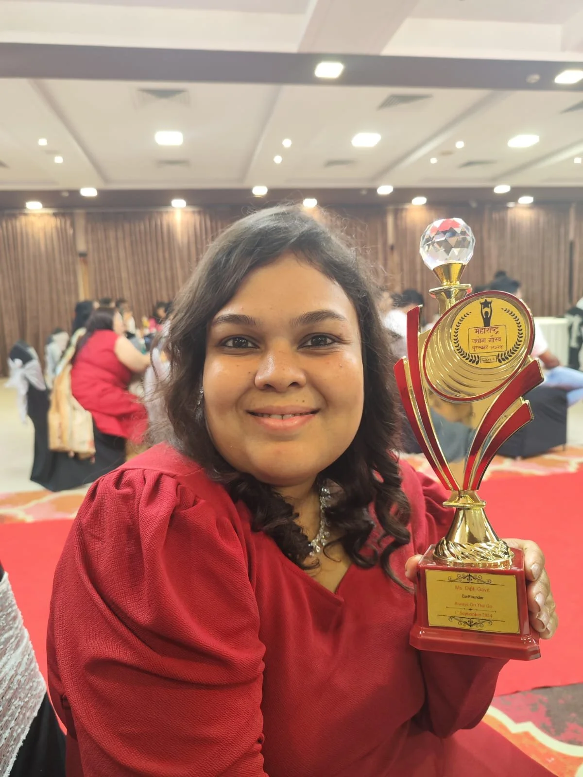 A woman smiling and holding a trophy at an indoor event, with others sitting and standing in the background.