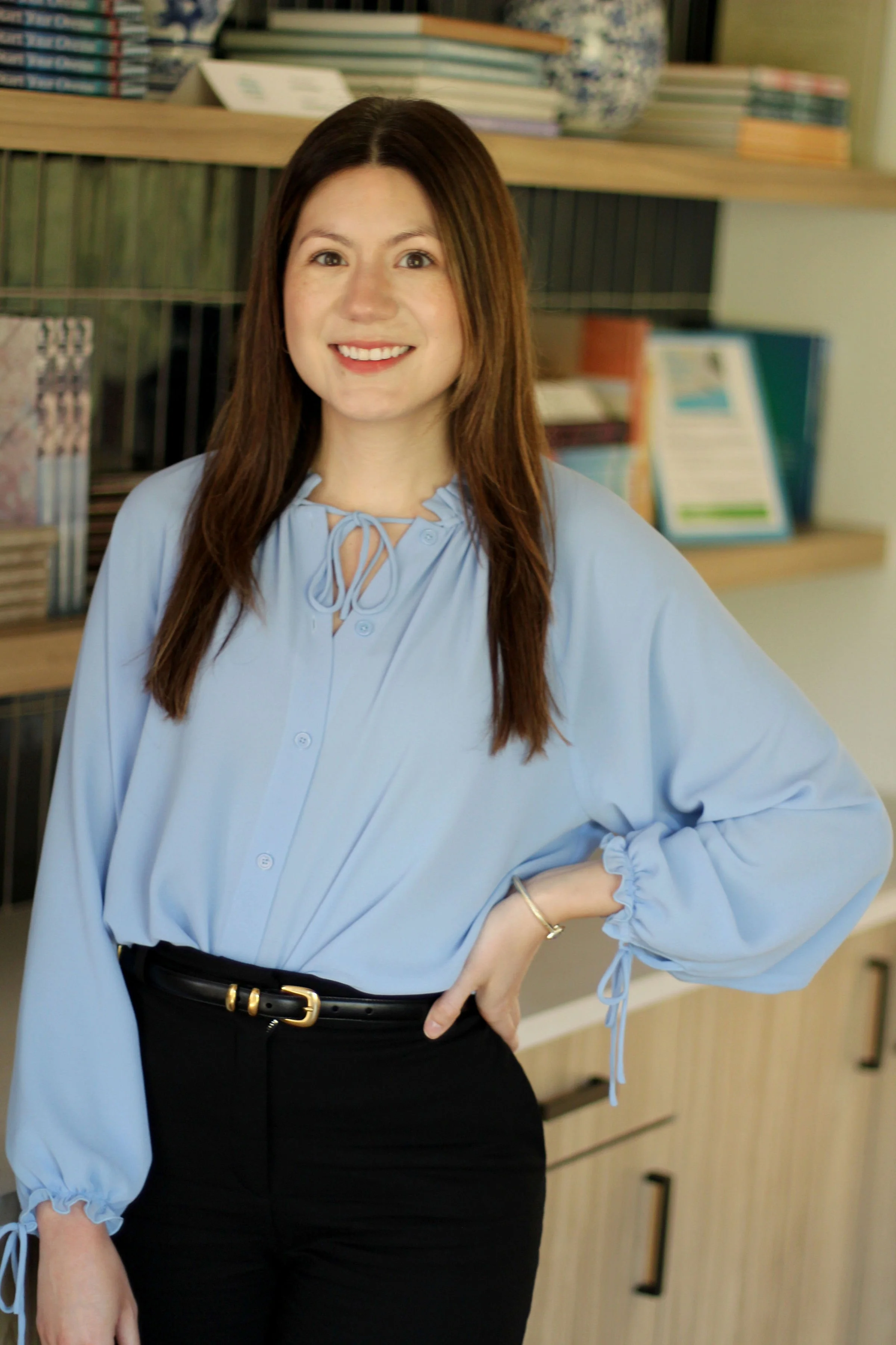 A woman with long brown hair smiling, wearing a light blue blouse with ruffled cuffs and black pants with a belt, standing in an office or library with shelves of books and binders in the background.