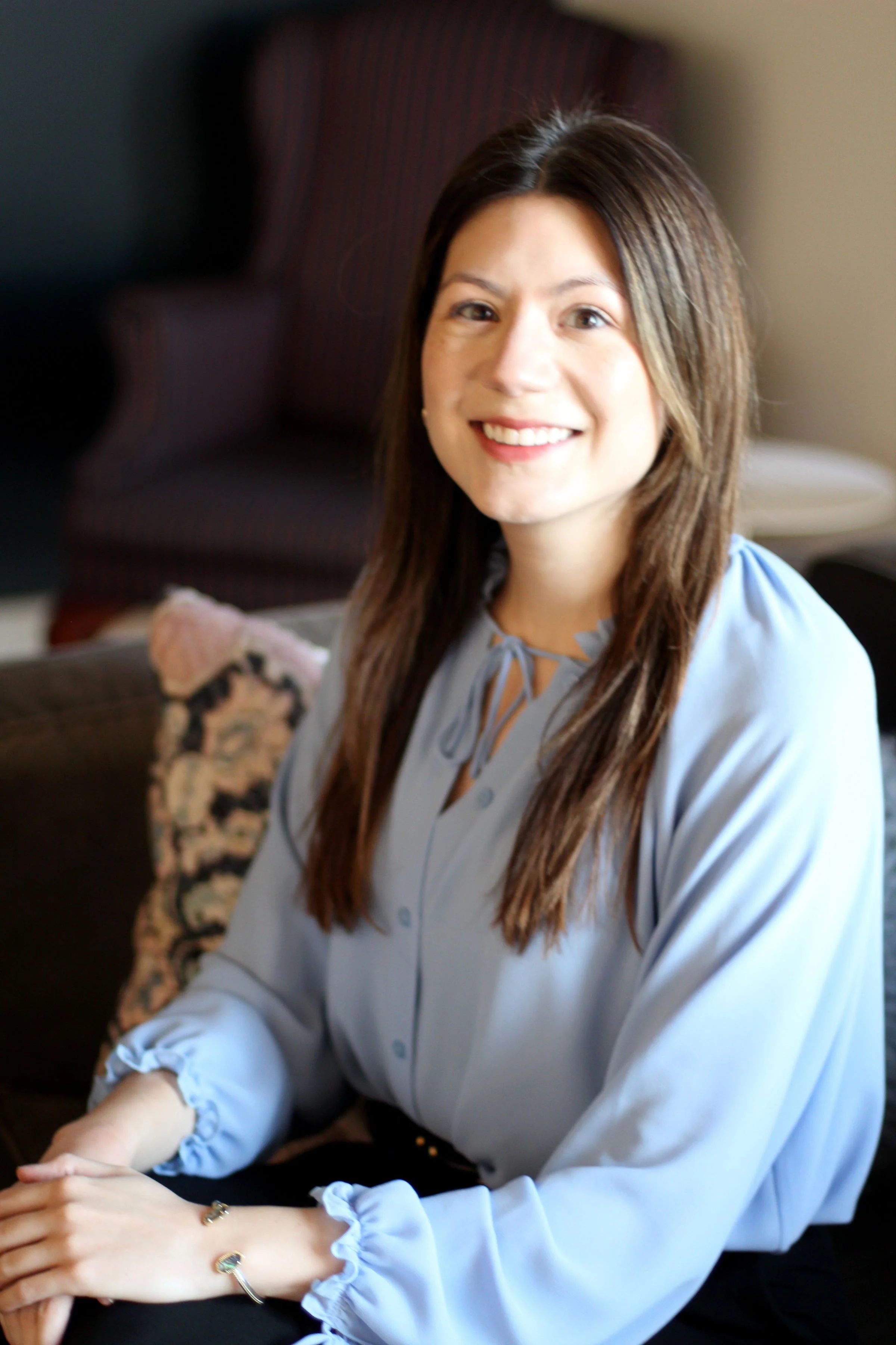 A woman smiling, with long brown hair, wearing a light blue blouse with ruffled cuffs, sitting at a table with her hands on her lap. There is a patterned pillow and a dark chair in the background.