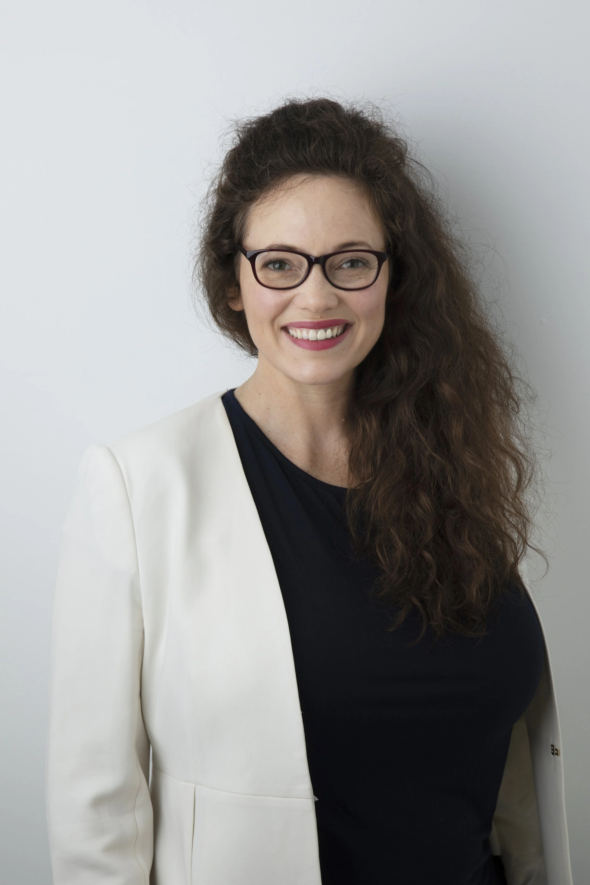A woman with long curly brown hair, wearing glasses, a navy blue top, and a white blazer, smiling against a plain white background.
