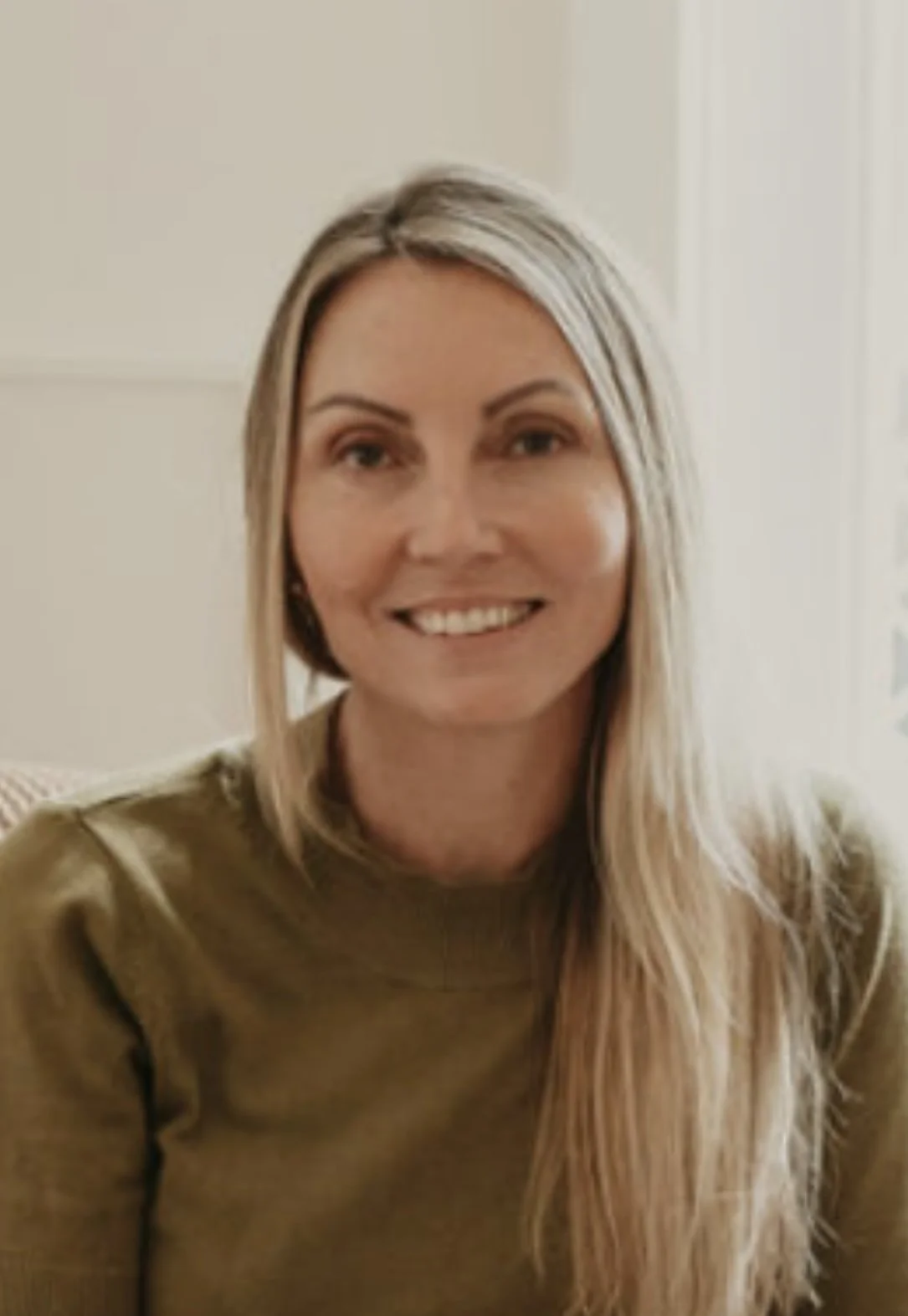 A woman with long blonde hair, smiling in a cozy indoor setting.