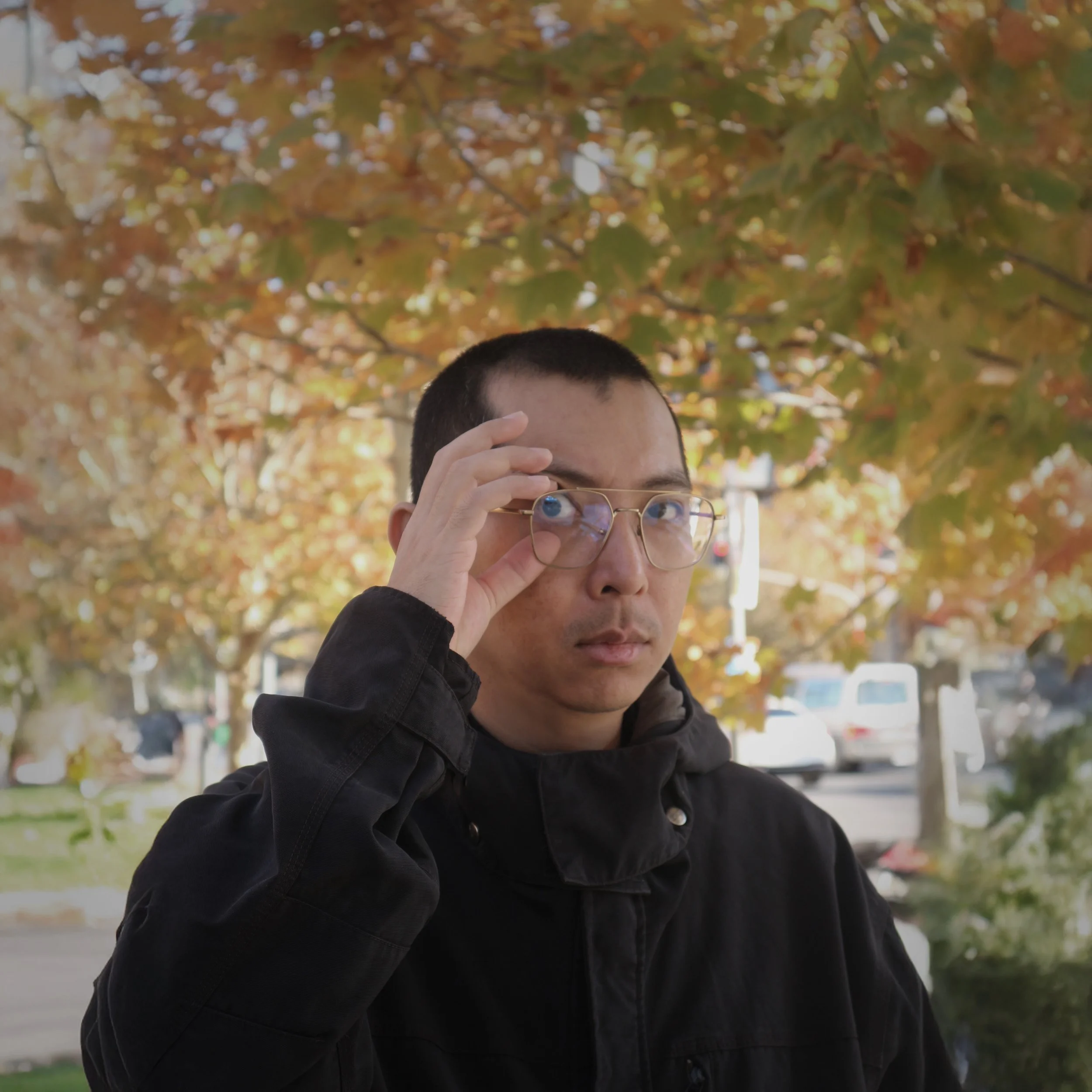 A young man with short black hair and glasses, wearing a black jacket, standing outdoors during fall with colorful leaves on trees in the background.