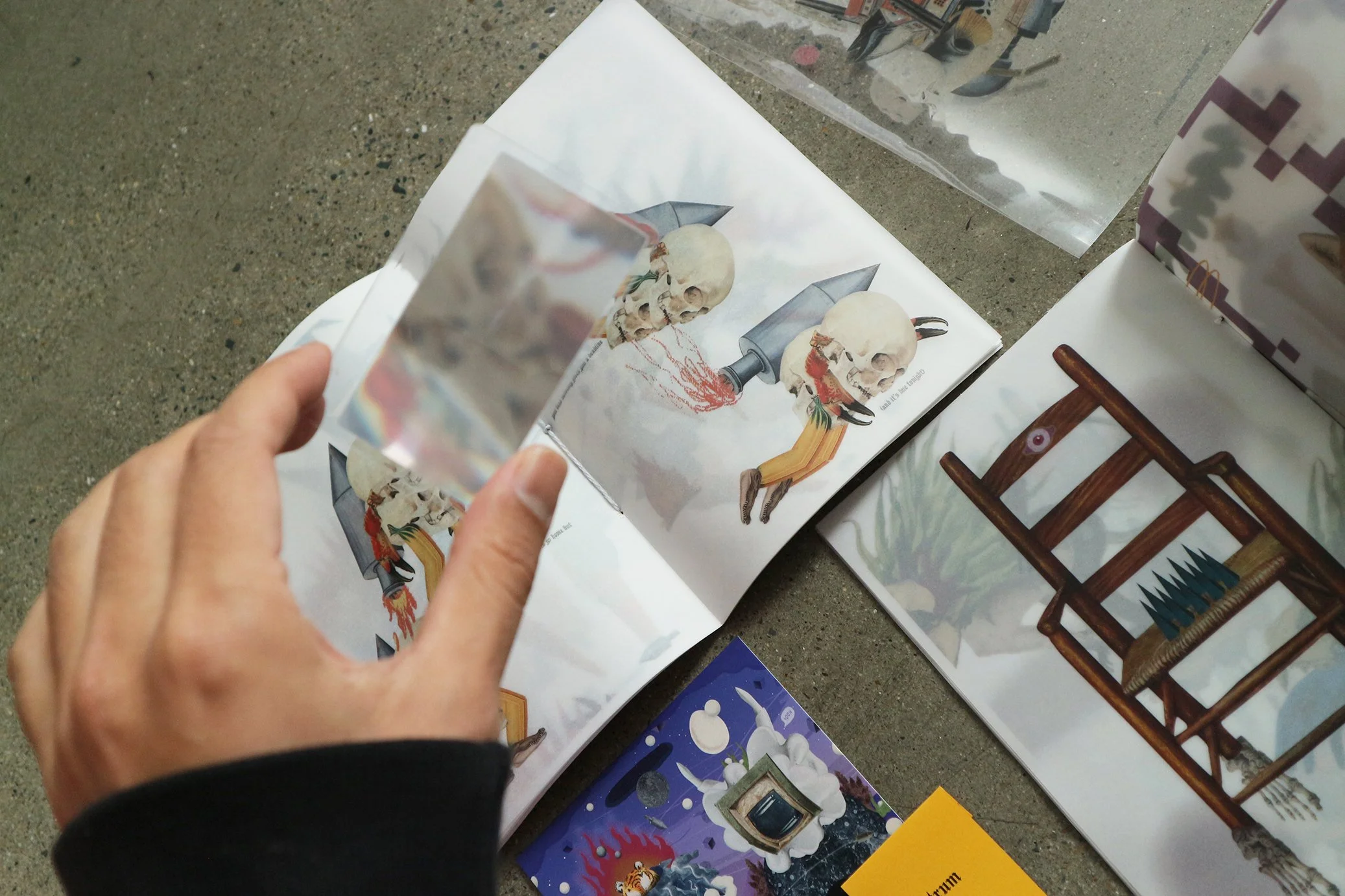A person holding a small card with an illustration of a skull and a gun, positioned over a larger poster of the same illustration in a classroom or office setting with various posters and items on a table.