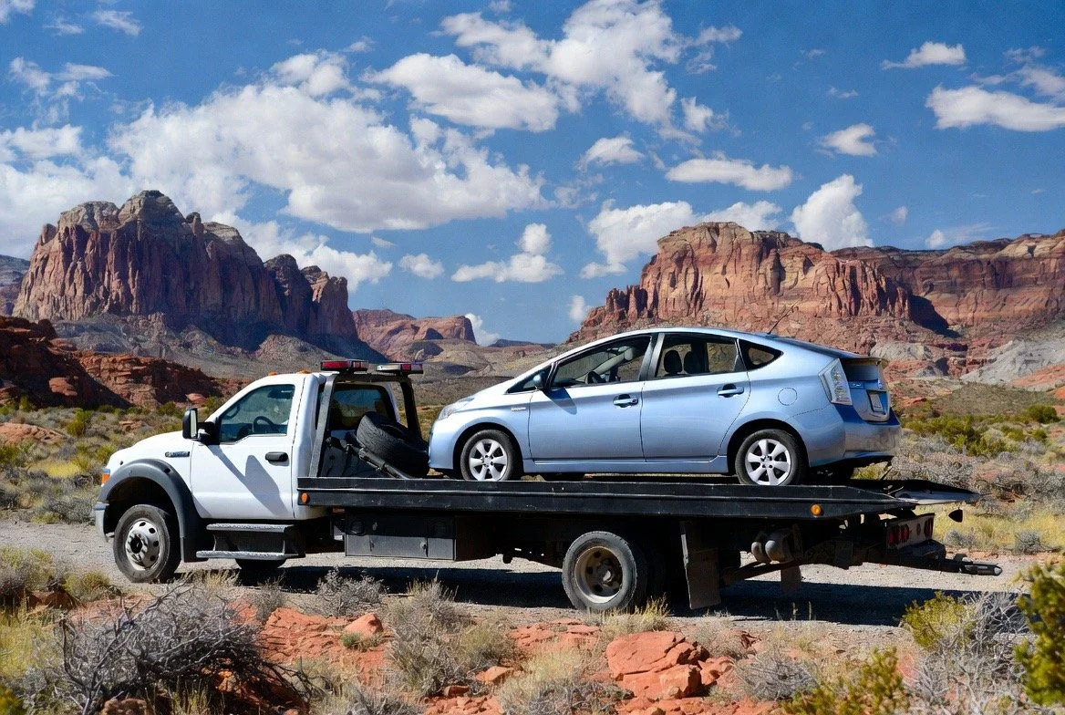 A tow truck carrying a blue sedan parked on a rural road near Ivins Utah with rock formations and mountains in the background under a partly cloudy sky.