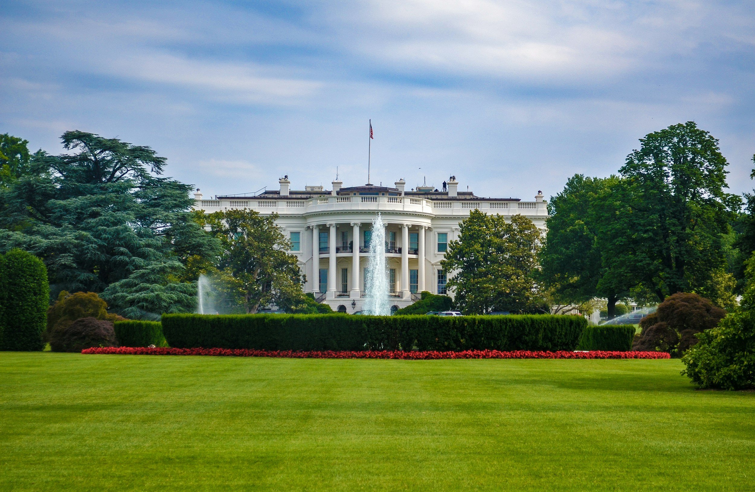 The White House, the official residence of the U.S. president, is visible behind trees and fountains, with a flag flying on the roof and a cloudy sky above.